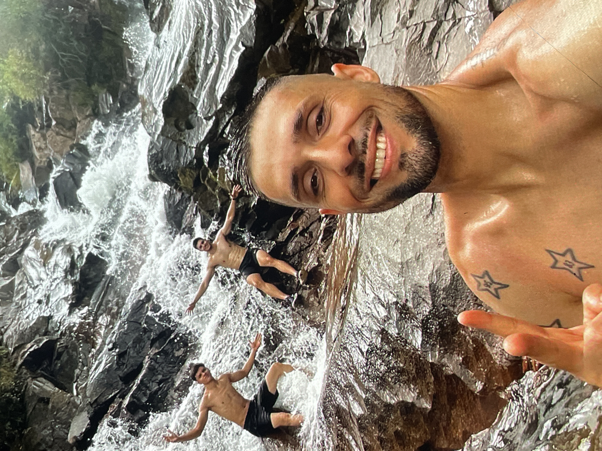 The image shows three men posing for a selfie in a natural waterfall environment, presumably near Harmanli, Bulgaria. The man taking the selfie is fair-skinned with a shaved head, a short beard, and noticeable tattoos of stars on his shoulder. He is smiling directly at the camera. Behind him and slightly below are two other men. Both of them are fair-skinned, shirtless, and wearing black shorts. They are lying amongst the rocks and water of the waterfall, with their arms outstretched, appearing to enjoy the moment. The environment consists of a rocky waterfall with water cascading over the rocks. There is some greenery visible on the left side of the frame.