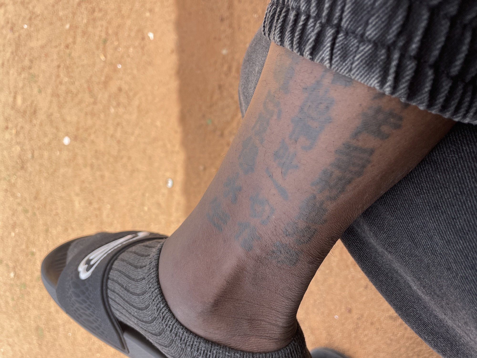 A top-down close-up shot depicts a person's lower leg and foot. The person is wearing gray socks, black Nike sandals, and gray denim pants. The leg has a faded, dark-colored tattoo featuring what appears to be Chinese or Japanese characters. The background is an out-of-focus ground surface, possibly dirt or sand. The exact location in the city of Jos, Nigeria, is not discernible from the image.