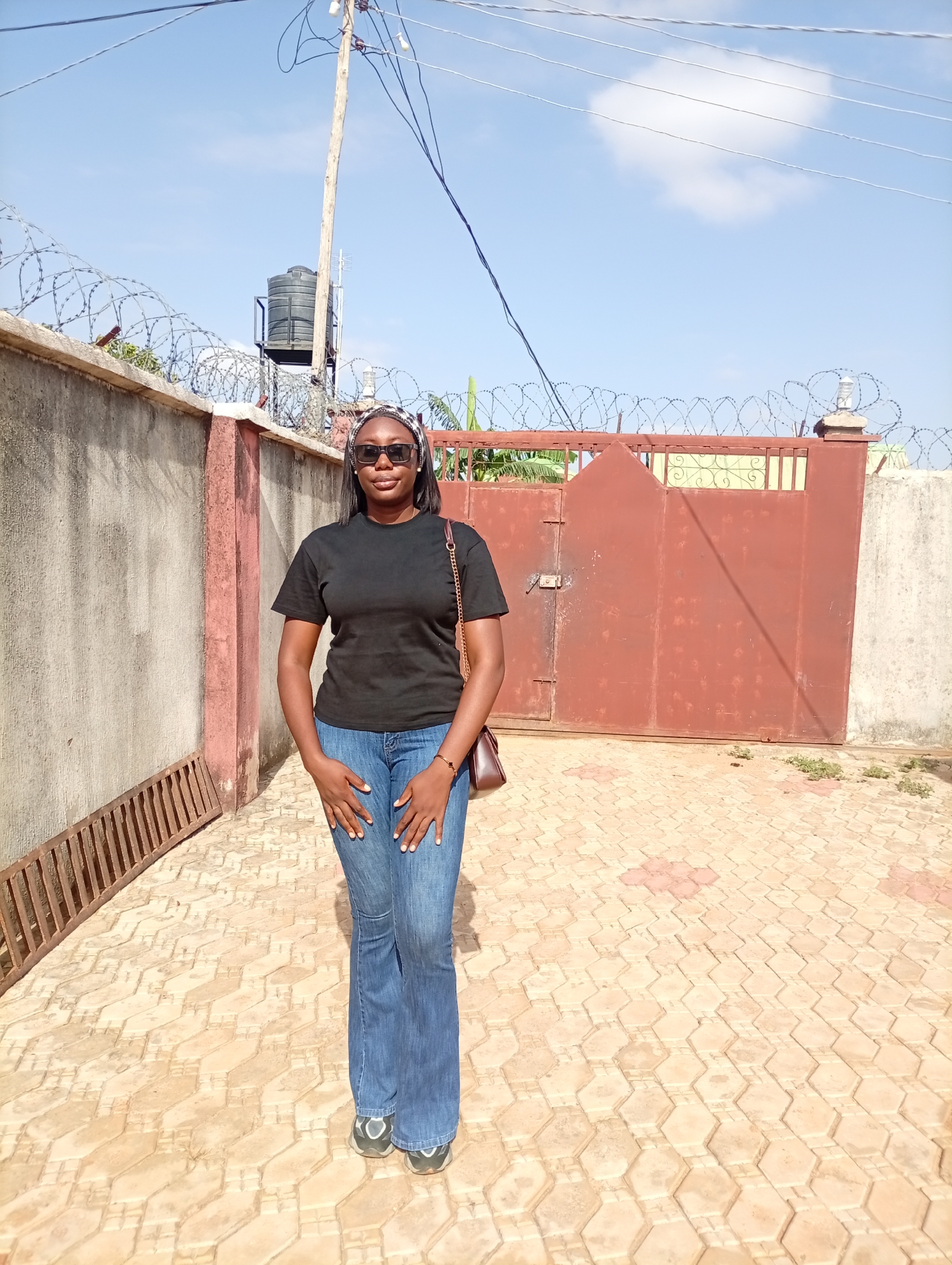 A woman stands centered in the frame, on a tan brick patio. She is wearing a black t-shirt, blue jeans, sunglasses, and a patterned headband. A small brown bag hangs from her right shoulder.
Behind the woman is a compound with concrete walls topped with barbed wire. A large, reddish-brown metal gate is visible in the center. A water tank sits on a metal frame behind the wall on the left. A wooden utility pole with wires is overhead.
The location is Rido, Nigeria.