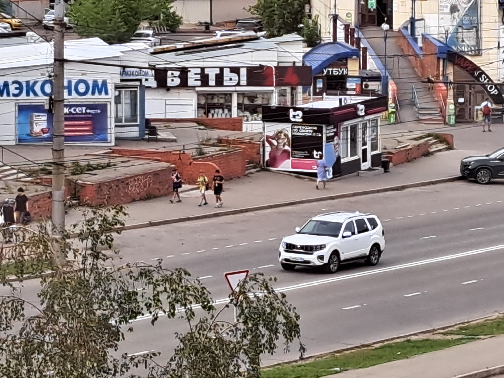 The image depicts an outdoor scene on a street in Irkutsk, Russia. In the foreground, a white SUV is driving on a paved road with visible lane markings. To the left of the road, a tree with green leaves partially obscures the view. Further back, a pedestrian walkway runs alongside the road, with several individuals walking. Beyond the walkway are buildings with commercial signage. One building displays the text "ЭКОНОМ" (Econom) and another shows "ВЕТЫ" (Vety). A small kiosk with advertisements is also present. In the background, there are more buildings and an elevated walkway with stairs, suggesting a complex urban environment. The overall impression is a snapshot of daily life in a commercial area of the city.