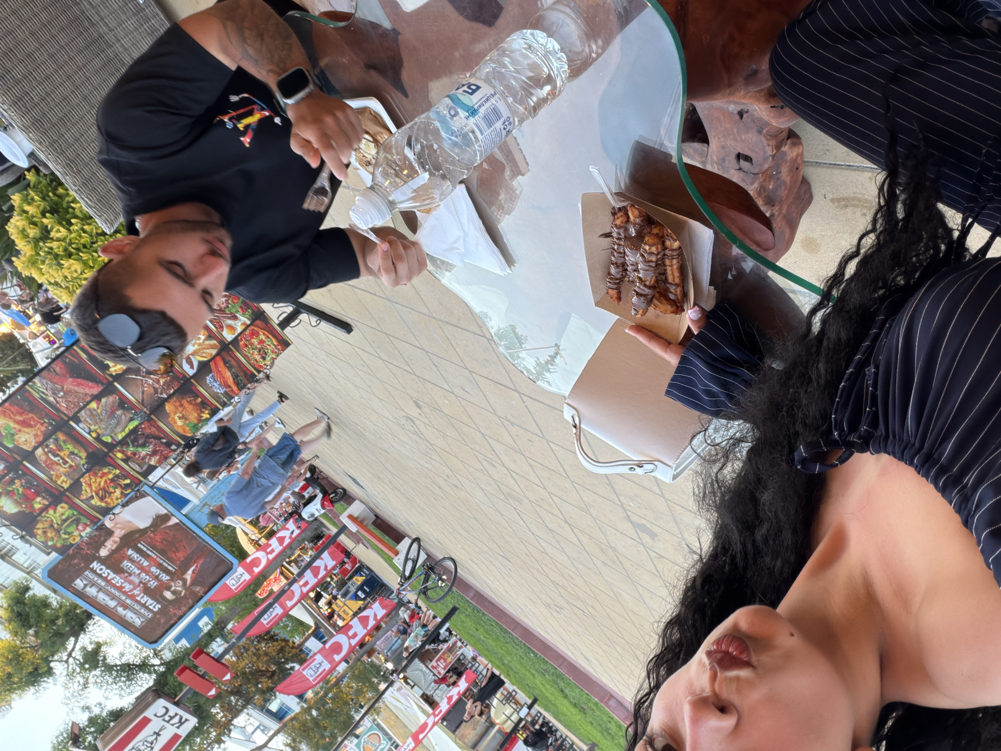 In Nesebar, Bulgaria, on a sunny day, a young woman with long, dark curly hair, wearing a navy blue striped off-the-shoulder top, leans forward and puckers her lips towards the camera. Beside her, a man wearing a black t-shirt and sunglasses is seated at a glass-topped table, holding a fork and about to eat from a plate of food. On the table, in addition to the man's plate and a bottle of water, is a small cardboard container filled with churros drizzled with chocolate. The background reveals a bustling outdoor scene with KFC branding, red flags, a billboard advertising an event, people walking by, a bicycle, and a scooter, all set against a backdrop of trees and buildings. The pavement is paved with diamond-shaped tiles.