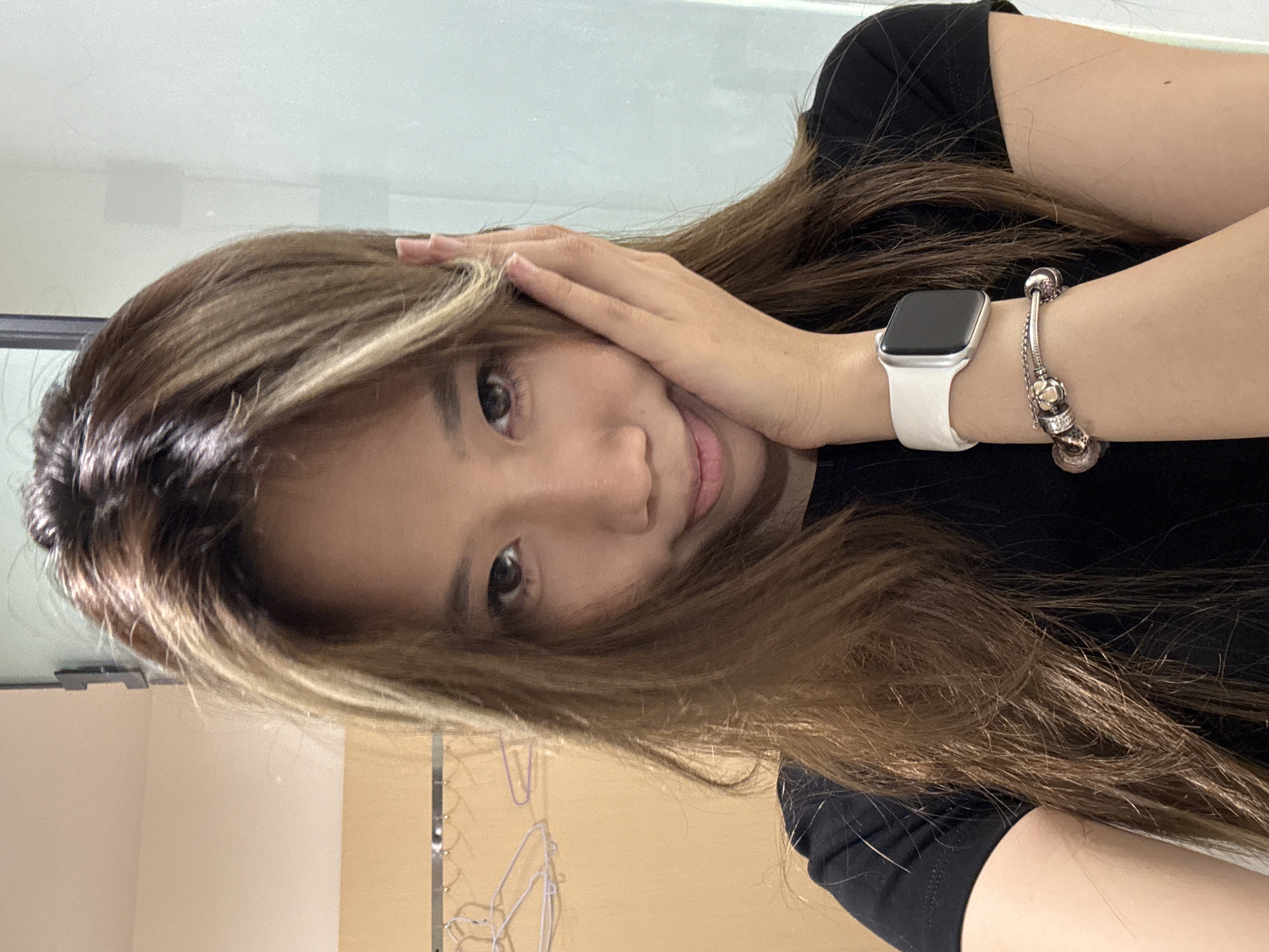 A young woman, likely of Asian descent, is featured in a close-up indoor portrait, with the image appearing tilted or taken at an angle, showing her head slightly to her left. She looks directly at the camera with a calm, pleasant expression and a subtle smile. Her long, straight hair, a mix of brown and lighter, possibly blonde or light brown highlights, frames her face and extends over her shoulders. Her left hand is gently placed near her temple, touching her hair.
She is wearing a black short-sleeved top. On her left wrist, she sports a silver-toned Apple Watch with a white band, its screen dark. Her right wrist is accessorized with a silver-colored charm bracelet adorned with multiple spherical charms.
The background is a simple indoor setting, likely a room in Yuzhong District, China. It features plain light-colored walls, possibly beige or white. In the lower left of the frame, hints of metal or plastic hangers are visible, suggesting a closet or drying area. A light, reflective surface, possibly a window or mirror, occupies the upper background. The lighting is soft and even, indicating an indoor environment with diffused light. There is no visible text in the image.