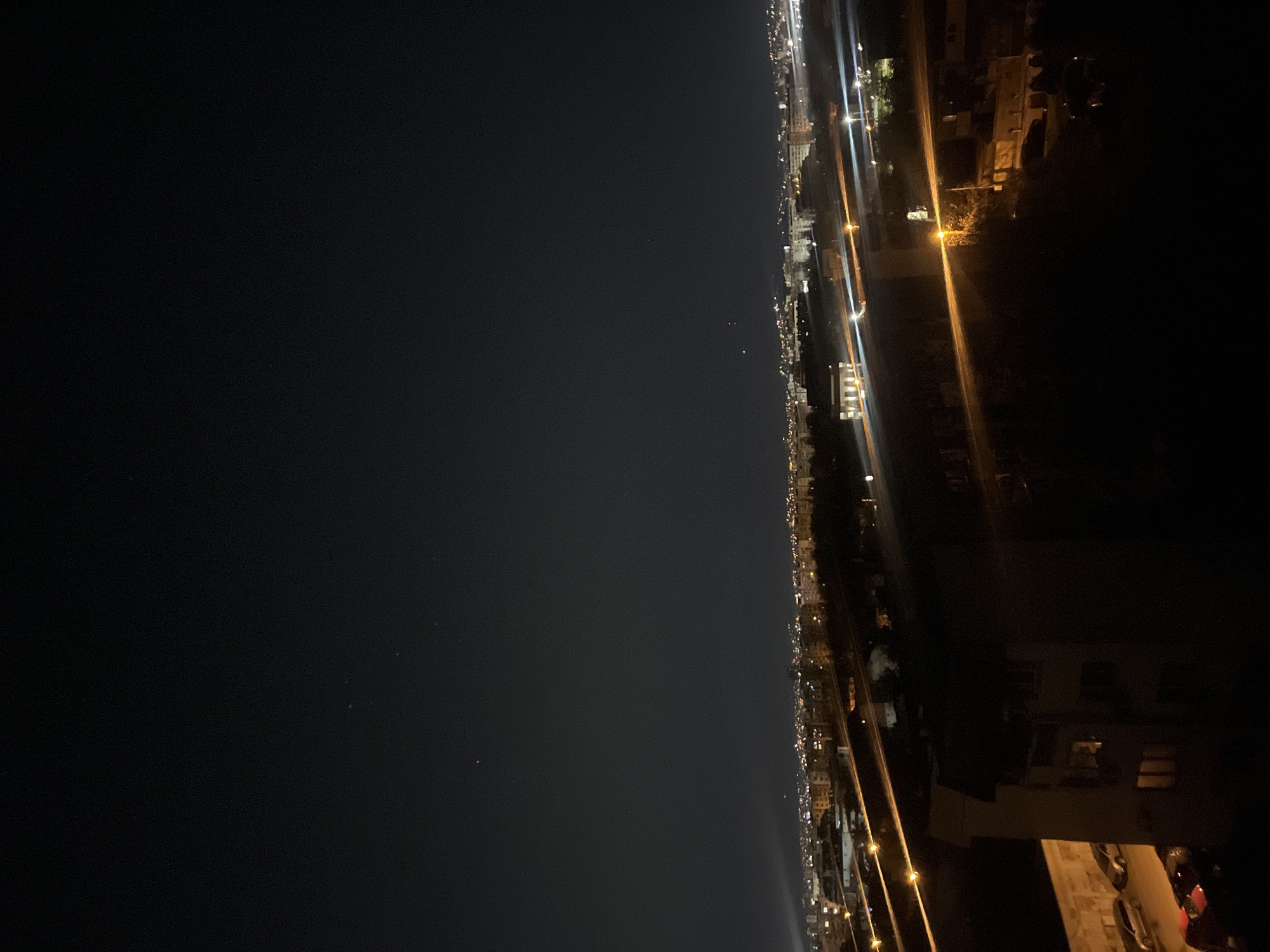 The image depicts a night view, likely taken from a high vantage point overlooking a city. On the right side of the image, urban structures, buildings, and roads illuminated by streetlights are visible. The left side of the image is mostly dark, potentially showing the night sky or a less illuminated part of the cityscape or body of water. The quality of the image suggests a long exposure time, as light sources appear elongated. I cannot verify the exact location in Sofia, Bulgaria.