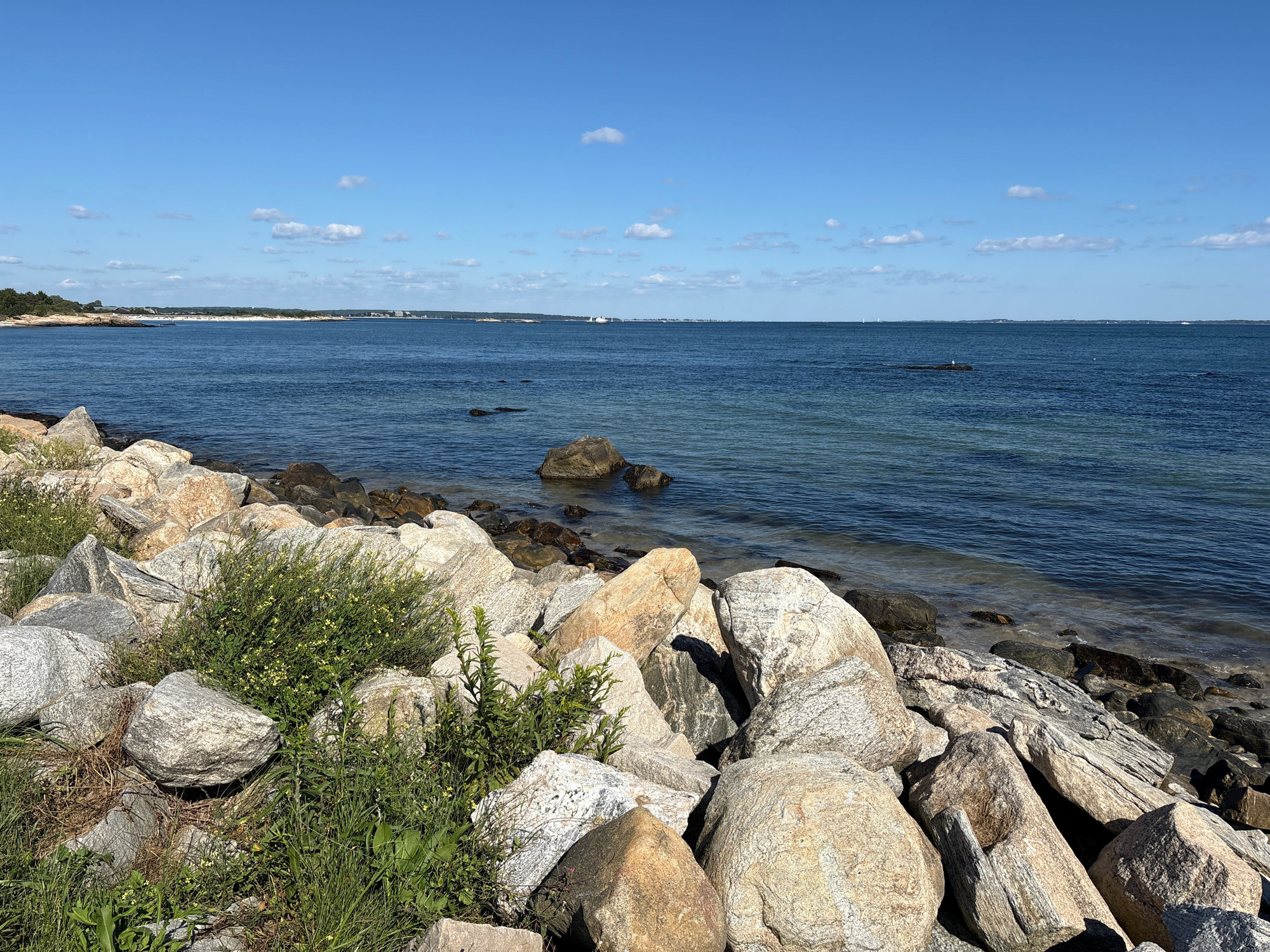 This is a wide, eye-level, outdoor photograph of a rocky shoreline on a clear, sunny day. The foreground is dominated by a jumble of large, light-colored boulders and smaller, darker rocks, with patches of green vegetation and a few small yellow wildflowers growing between them. The middle ground consists of the calm, blue ocean, with several dark rocks partially submerged and a distant, small white buoy visible. The horizon line is low, with a sliver of land on the far left featuring trees and structures, and the vast expanse of the blue sky, dotted with a few small white clouds, taking up the upper portion of the image. The scene appears to be a natural coastline, possibly in Groton, Connecticut, United States of America, with no people or specific human activity depicted.