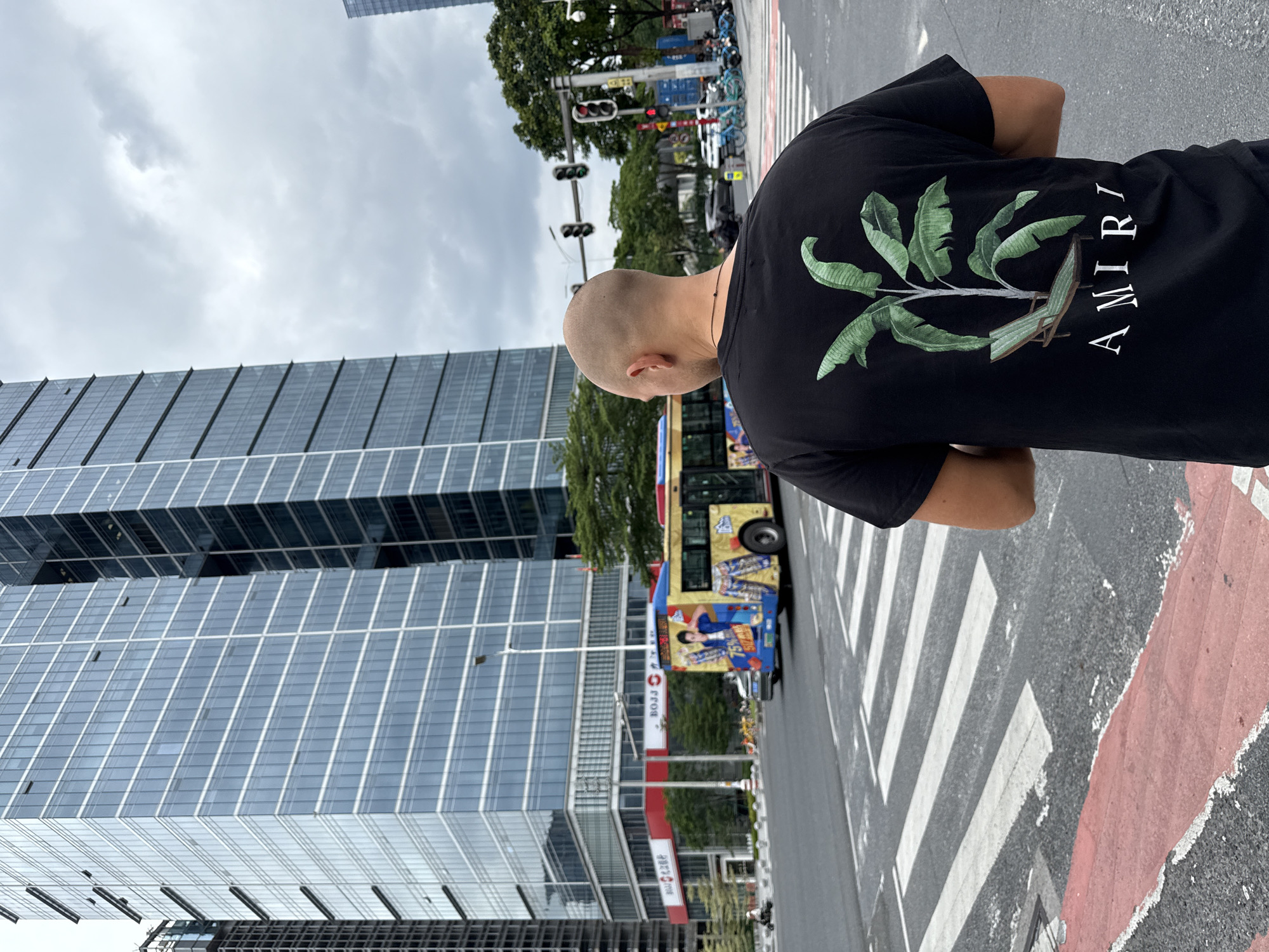 A bald man wearing a black t-shirt with a banana leaf and chaise lounge graphic stands on a crosswalk in Tianhe District, China, facing away from the camera. The t-shirt also has the word "AMIRI" printed in white below the graphic. In the background, a yellow bus with graphics of people and pants drives past tall, modern glass buildings and traffic lights. The sky is overcast.