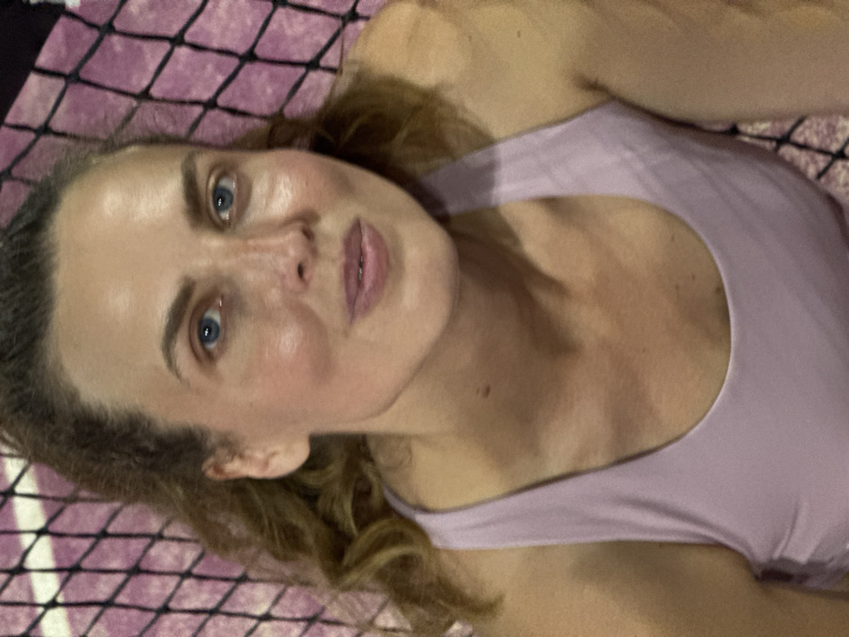 The image presents a close-up, eye-level portrait of an adult female located in Pecatu, Indonesia. She possesses light brown or auburn hair, which appears to be pulled back, with wavy strands visible on the left side of her face. Her eyes are blue, and her complexion is fair, showing a slight flush on the cheeks and forehead. She is wearing a light purple tank top. Her facial expression is neutral, and she is looking directly into the camera. The background is out of focus, displaying a dark grid pattern over a light purple or pinkish surface, suggestive of a net or a woven textured material, indicating she is potentially reclining or positioned against such a surface.