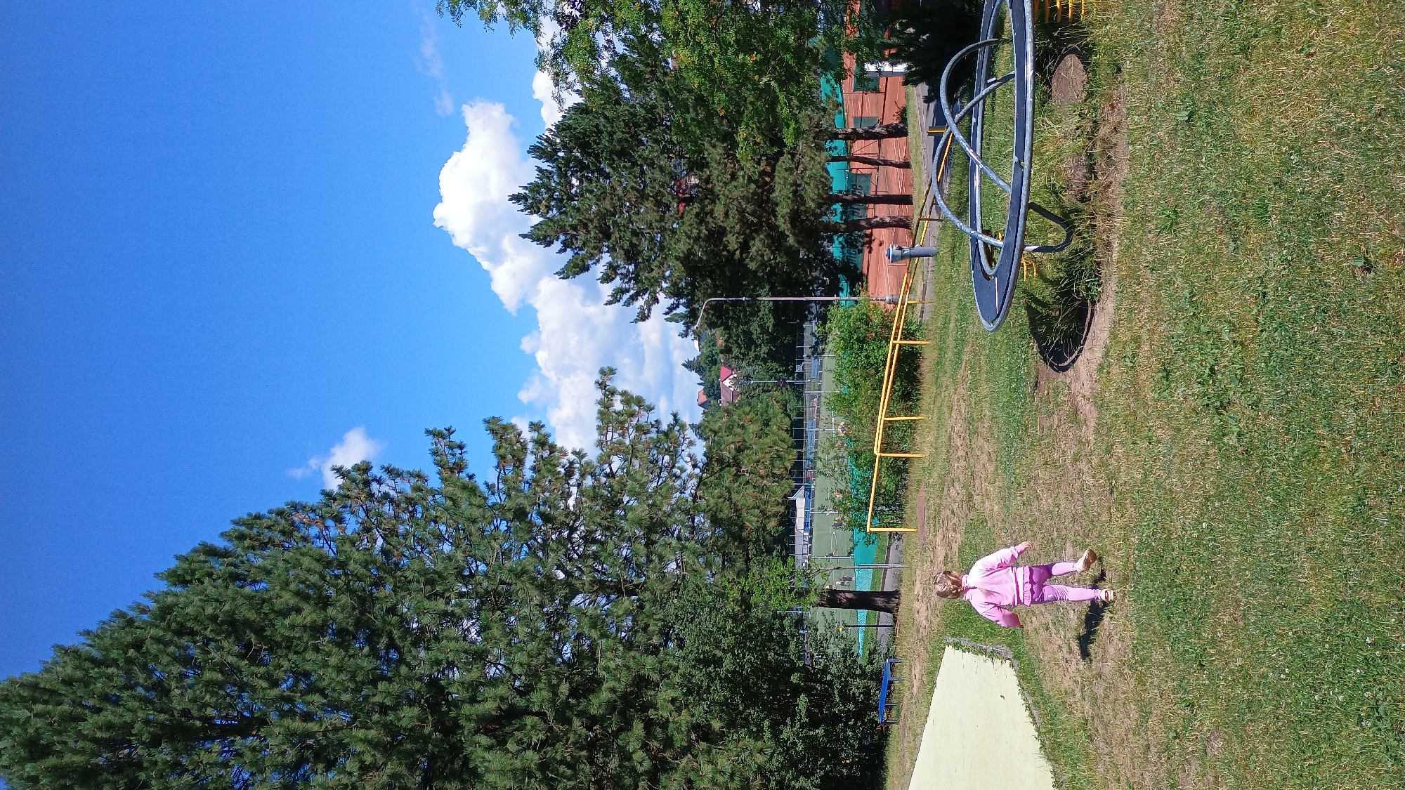 A young child with blonde hair in a braid, wearing a pink tracksuit, walks away from the camera across a grassy area in what appears to be a park or playground. The child is moving towards a black and silver merry-go-round. In the background, there are trees, a fence, a sports field, tennis courts with red clay surfaces, and a few houses with red roofs on a distant hill. The sky is bright blue with scattered white clouds. The scene is illuminated by daylight, suggesting it is a clear, sunny day. The location is the Sokolov Municipal Stadium sports complex in Sokolov, Czechia, specifically the area between the stadium and the tennis courts.