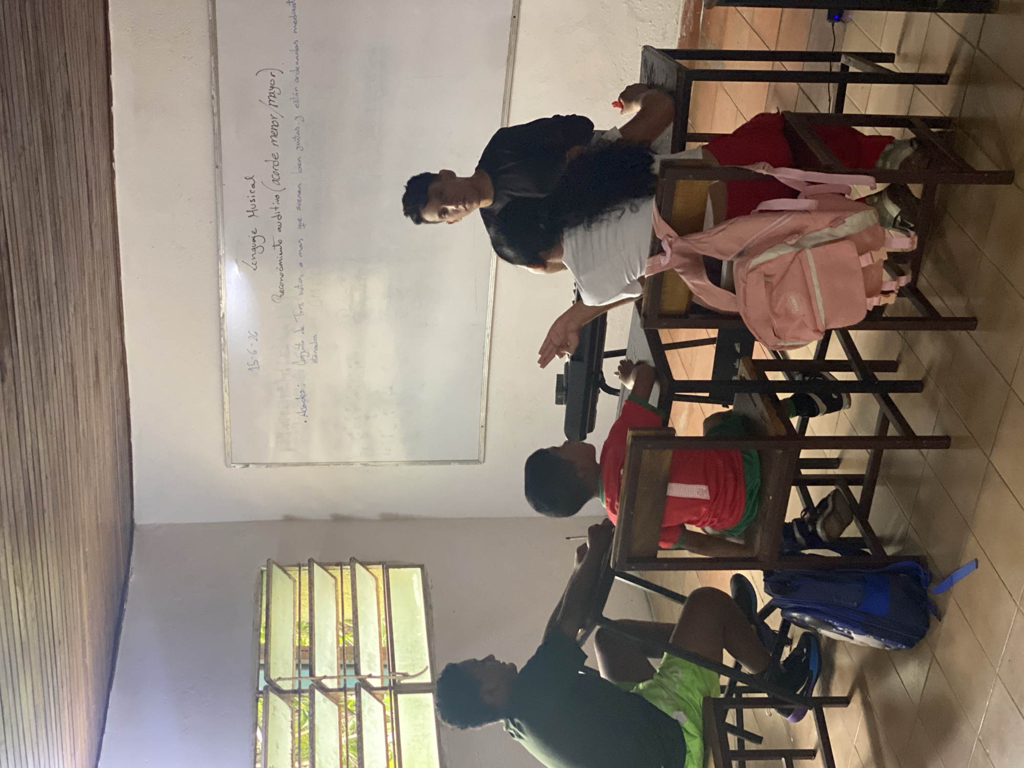 A group of students are gathered in a classroom, with a teacher standing at the front. The classroom appears to be a modest, possibly rural, setting. A whiteboard is visible behind the teacher, with some handwritten notes about "Lenguaje Musical" and "Reconocimiento auditivo (acorde menor/mayor)". There are multiple desks and chairs, with some students seated and others standing. One student, wearing a black shirt, stands and gestures towards the whiteboard, while another student, wearing a white shirt and a pink backpack, sits facing away from the camera with their hand raised. Two other students, one wearing a green t-shirt and shorts, and another in a red shirt, are also seated at desks. The room has large windows with louvers, allowing natural light to enter. The ceiling is made of wood panels. The floor is tiled. The overall impression is of an educational setting, likely a school, possibly in Tinaquillo, Venezuela, given the context.