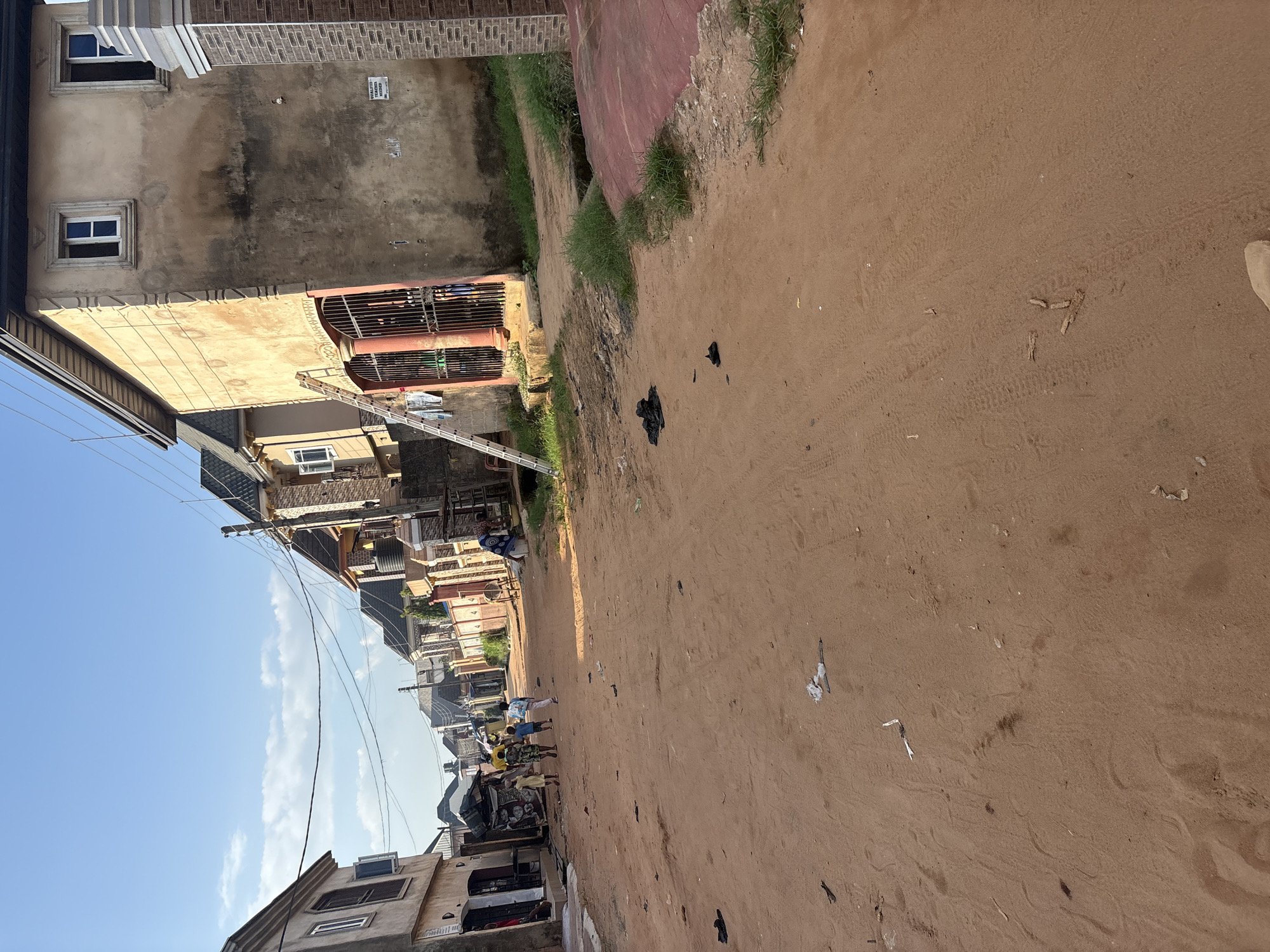 The image depicts a dusty, unpaved street in Obosi, Nigeria, lined with residential buildings. The sky is clear with scattered clouds, indicating daytime. The street is covered in reddish-brown sand and shows tire tracks. Several buildings are visible, some appearing to be under construction or in a state of disrepair, with exposed walls and unfinished elements. One building features a metallic gate and a ladder leaning against it, with laundry hanging on the ladder. There are a few people on the street, including children walking and a person sitting near a building. Scattered debris, including plastic bags and other refuse, is visible on the ground. The overall scene suggests a typical street in a developing urban area.