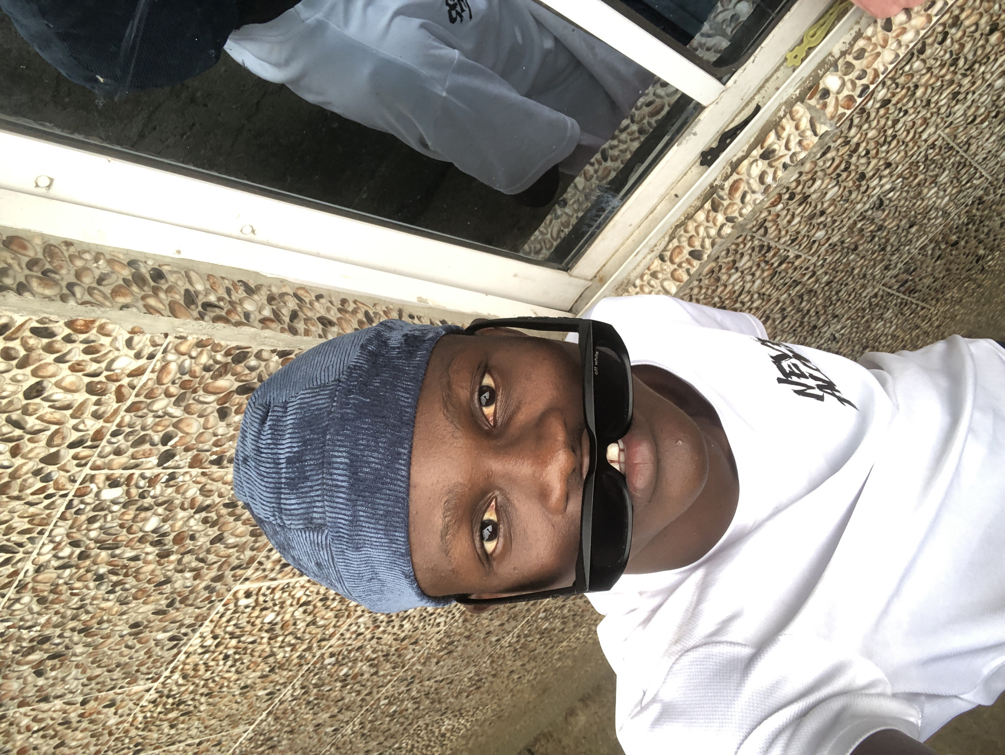 A medium shot captures a young Black person from the chest up, facing the camera. They wear a blue ribbed beanie pulled low on their forehead and a white t-shirt with a black graphic logo on the left chest. A pair of black sunglasses is perched on their nose, slightly obscuring their mouth. The background features a textured wall made of small, rounded pebbles in shades of brown and beige, juxtaposed with a white window frame and reflective glass. The overall setting suggests an outdoor location, possibly in Ado, Nigeria, given the architectural style and ambient light.