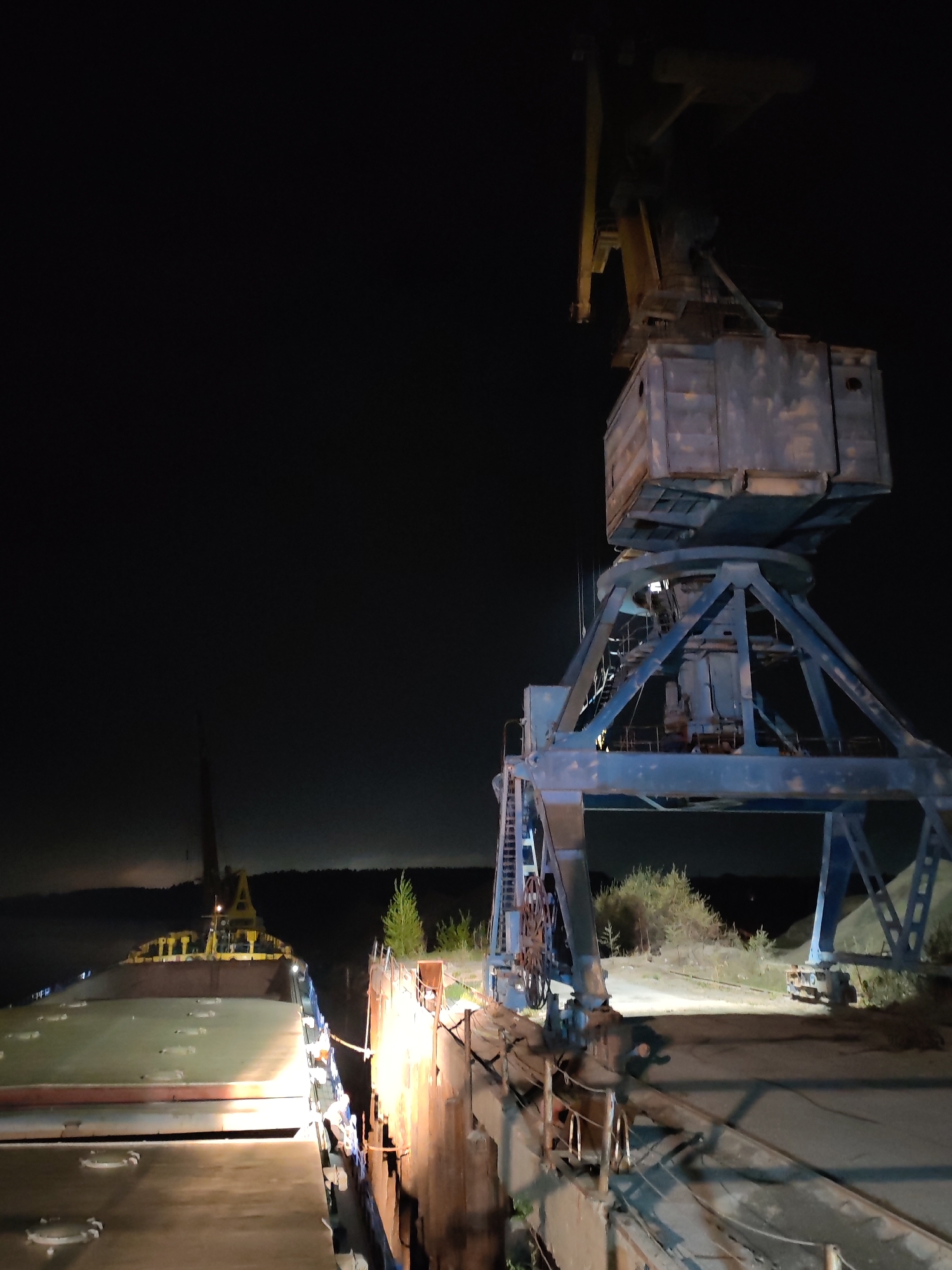 The image depicts a waterfront scene at night in Tver, Russia. A large, blue industrial crane dominates the right side of the frame. To the left, a section of a barge is visible, its flat deck illuminated by a light source. The barge is moored alongside a weathered concrete dock. The background is dark, obscuring details, but a line of trees is faintly visible on the horizon. The lighting conditions suggest the primary light sources are artificial, illuminating the barge and the crane. The scene appears to be in an industrial or commercial area, possibly a port or shipping facility.