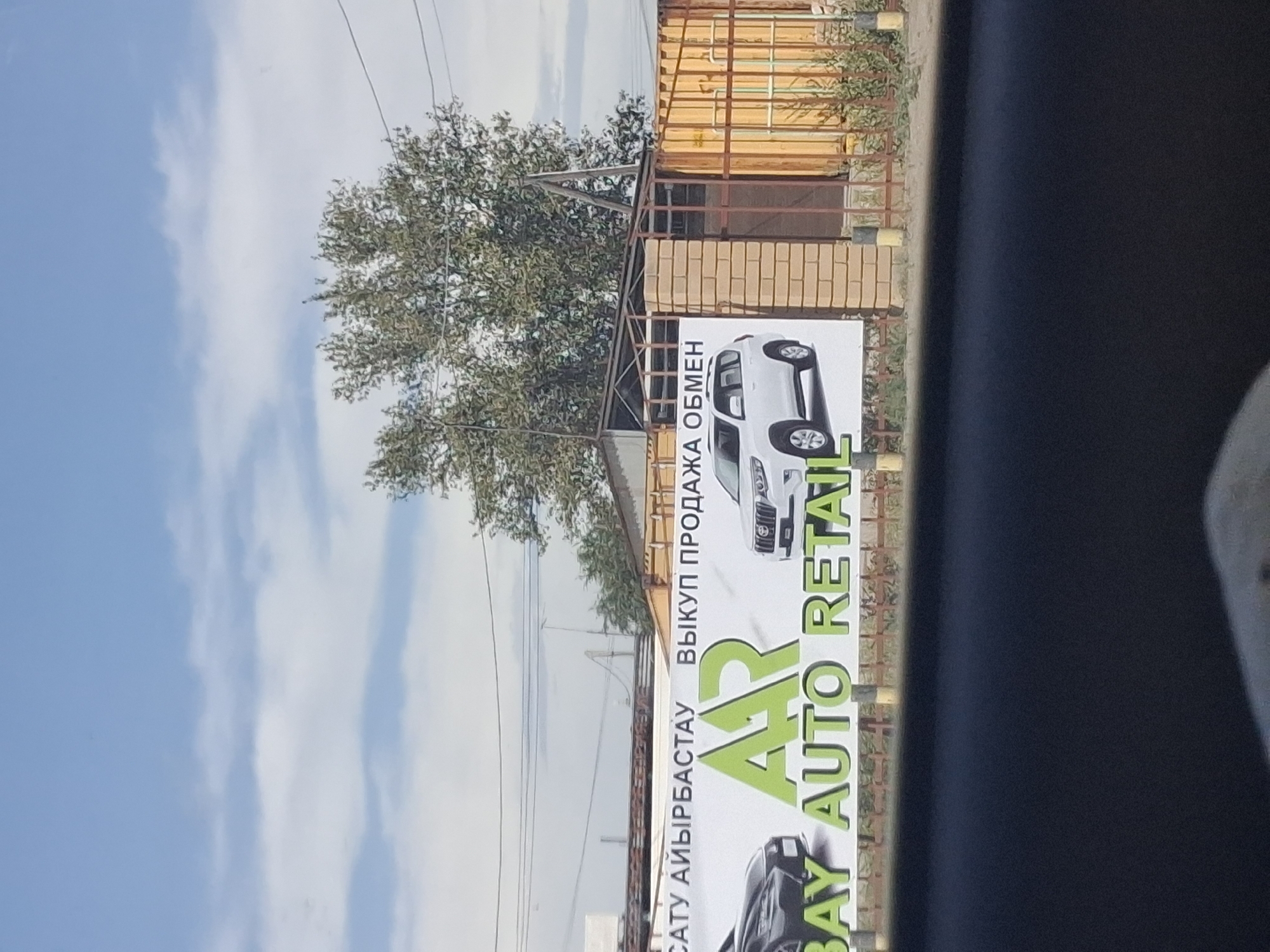 The image shows a banner for "AAR BAY AUTO RETAIL" in Semey, Kazakhstan. The banner features images of two cars, one black and one white SUV. Text on the banner appears to be in both Kazakh and Russian. The background includes a building with a brick pillar, a metal fence, and trees. The sky is partly cloudy. The perspective suggests the photo was taken from a vehicle.