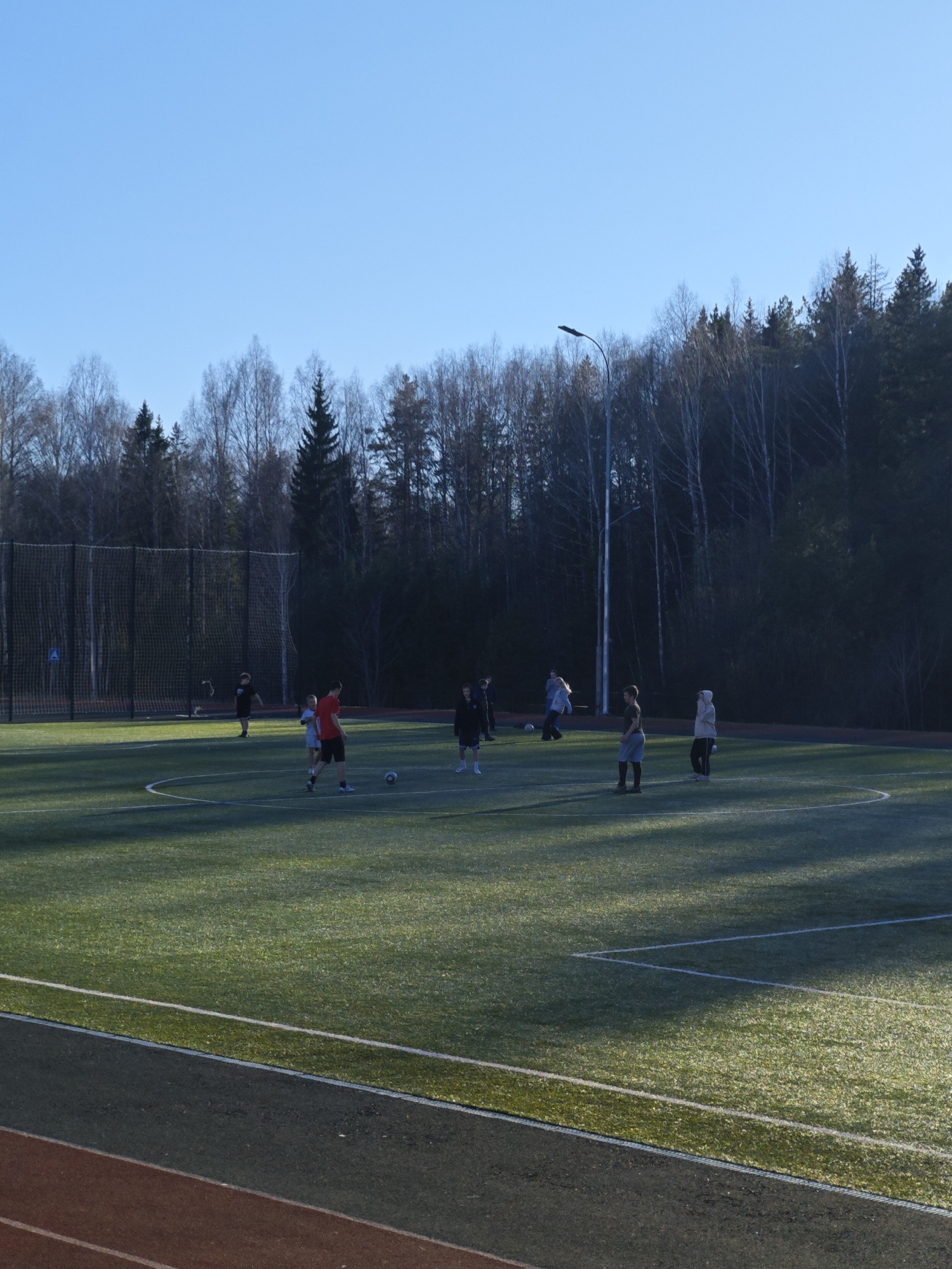 A group of people are playing soccer on a green turf field in Markovskii, Russia. The field is surrounded by a running track and trees. The sky is blue and there is a streetlight in the background. Some people are walking, while others are actively involved in the game. A net is visible to the left of the field.