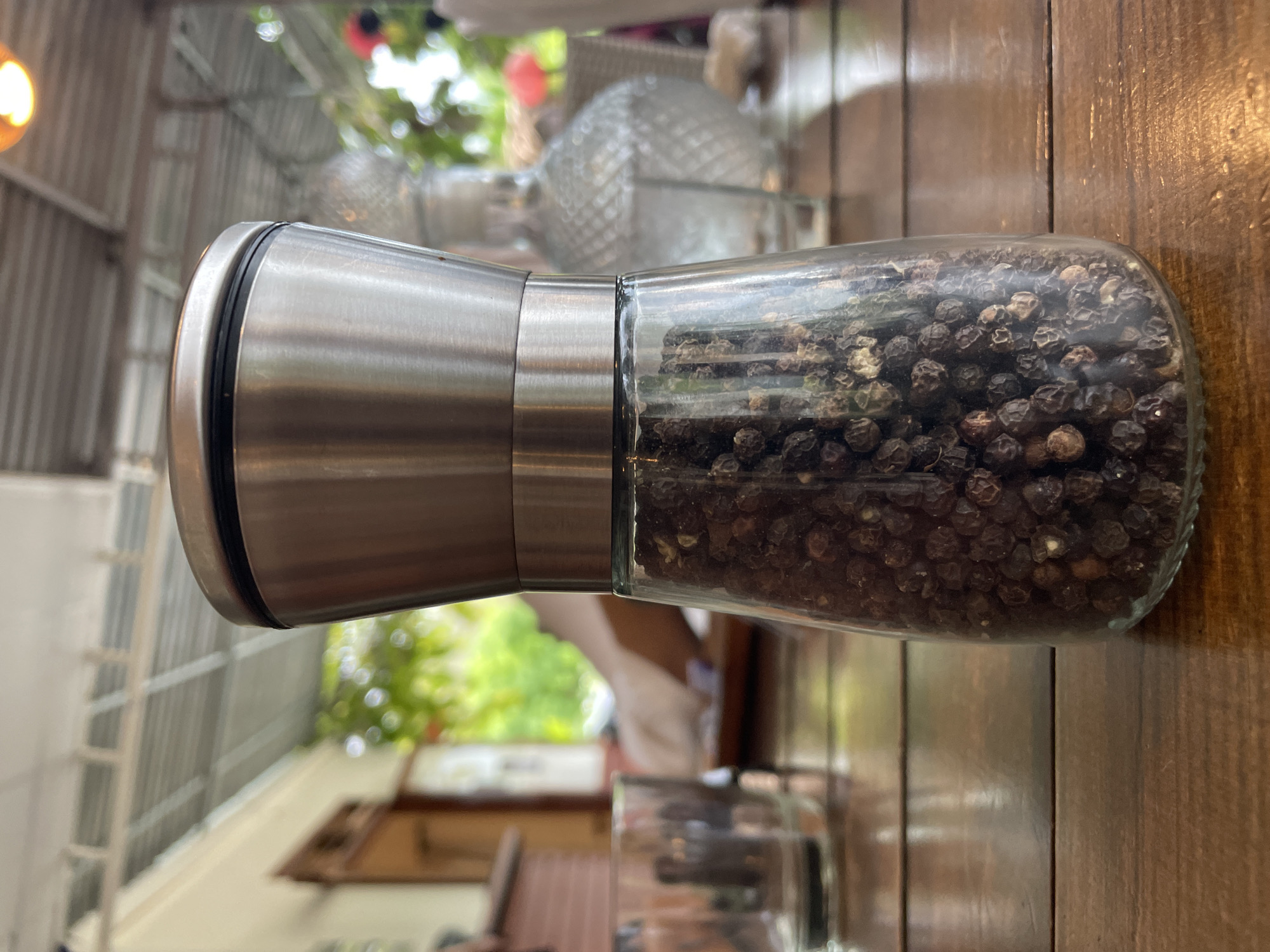 A pepper grinder is on a wooden table in Bac Nha Trang Ward, Vietnam. The grinder has a brushed silver metal top, a clear glass middle section filled with black peppercorns, and is lying horizontally. A blurred background shows another glass bottle and outdoor scenery. The lighting is bright.
