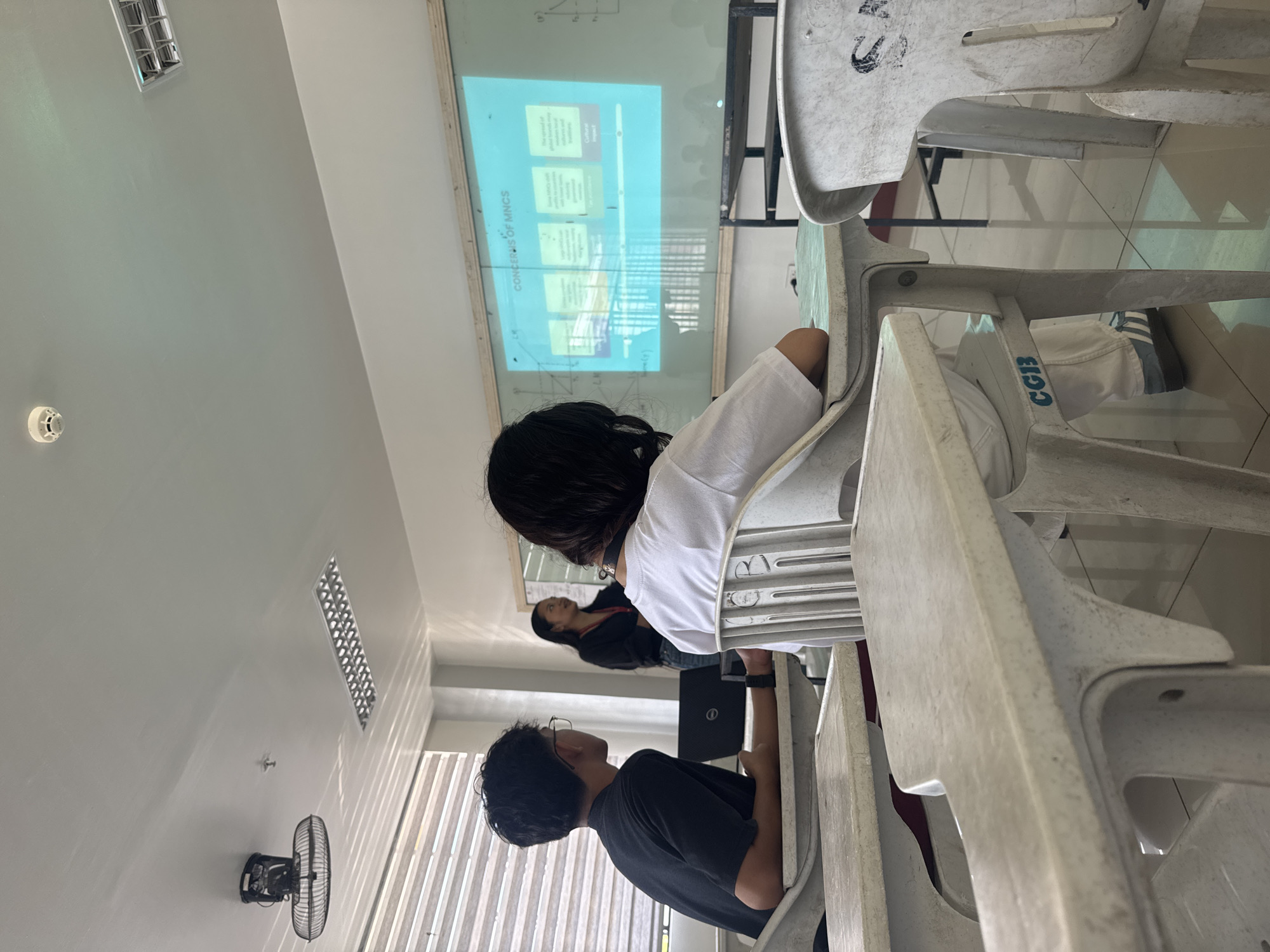 The image depicts a classroom setting in Davao City, Philippines. There are three individuals present: one in a white shirt, one in a black shirt, and another with dark hair wearing a black top. The people are seated in white plastic chairs, facing a projector screen displaying text and diagrams, likely a lecture on "Concerns of MNCs." A black laptop is visible in the scene. A wall-mounted fan is present near a window with blinds. The setting appears to be a typical indoor learning environment.