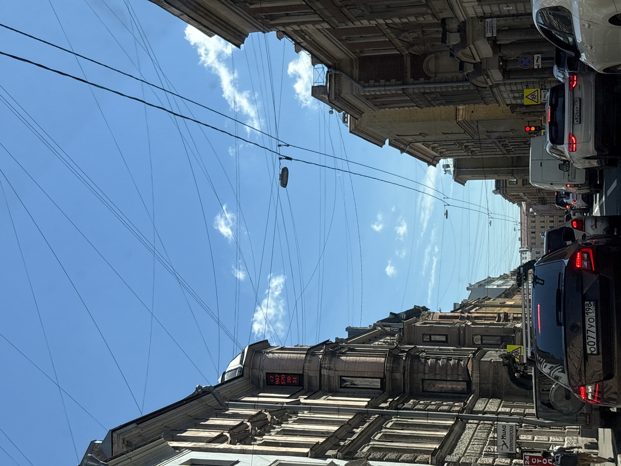 The image depicts a traffic jam on a city street in Saint Petersburg, Russia, under a clear blue sky with scattered clouds. The perspective is from a vehicle looking forward. On the left side of the street, a multi-story building with ornate architecture and windows is visible. A digital display on the building shows red numbers, potentially indicating temperature or time. Further down the street, smaller buildings and more complex architectural details can be seen.
On the right side, another multi-story building with similar classical architectural features lines the street. The street is filled with various vehicles, including cars, a van, and possibly a larger truck. Traffic lights are visible, with one showing red and amber. Street signs and utility poles with numerous overhead wires crisscross the sky, a common sight in older urban areas.
The foreground is dominated by the back of a dark-colored car with its license plate visible. To its left, a lighter-colored SUV is also present. A person with a backpack is walking on the sidewalk on the left side of the street, heading away from the viewer. The overall impression is a typical urban scene experiencing traffic congestion on a sunny day.