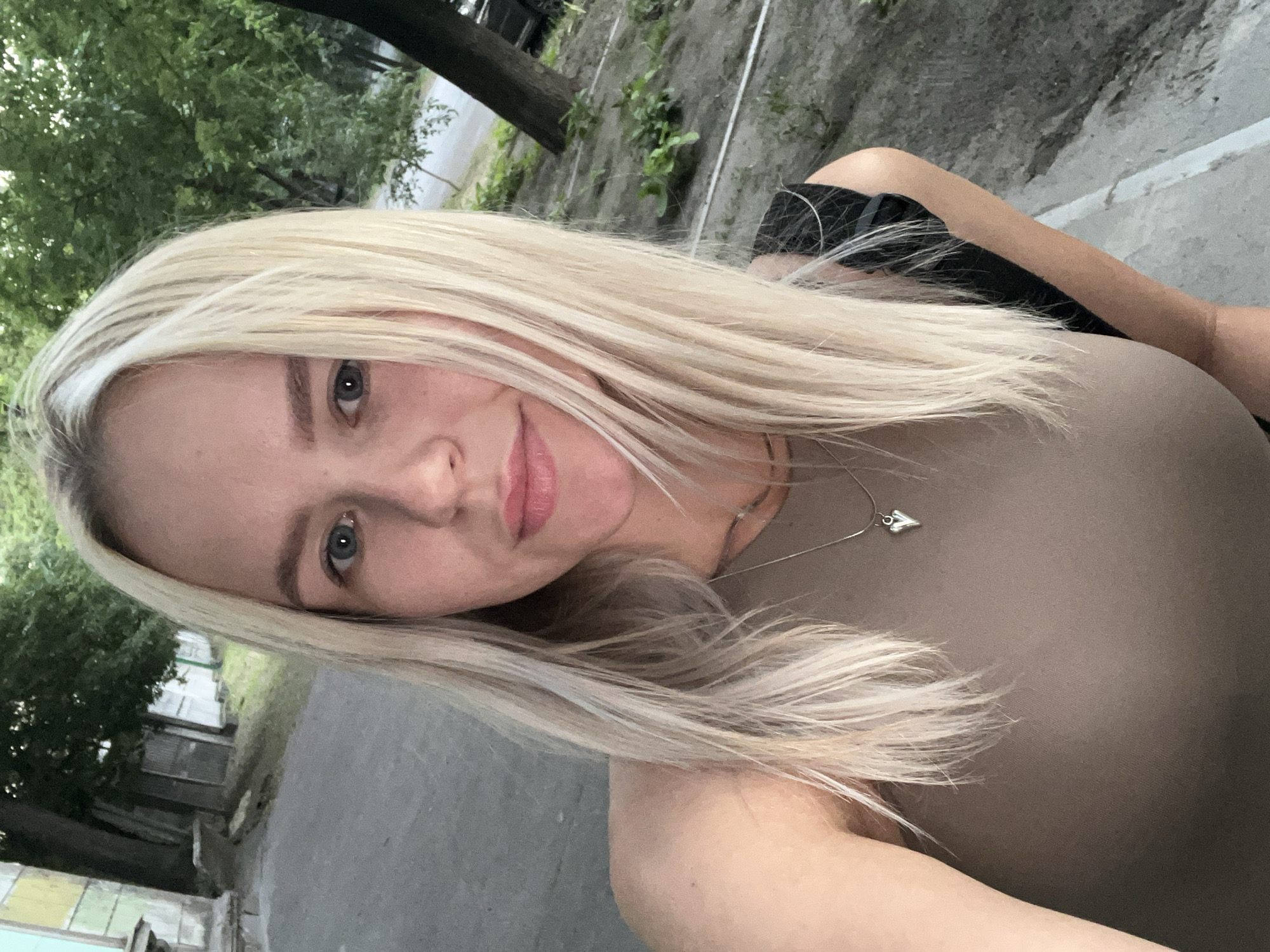 A medium shot selfie captures a young Caucasian woman with blonde hair and blue eyes, looking directly at the camera. She is wearing a taupe sleeveless top and a silver necklace with a heart-shaped pendant. Her hair is long and straight, falling loosely around her face and shoulders. The background appears to be an outdoor urban setting, possibly a street or park, with trees and some buildings visible. The lighting suggests it is daytime, though the overall tone is somewhat muted.