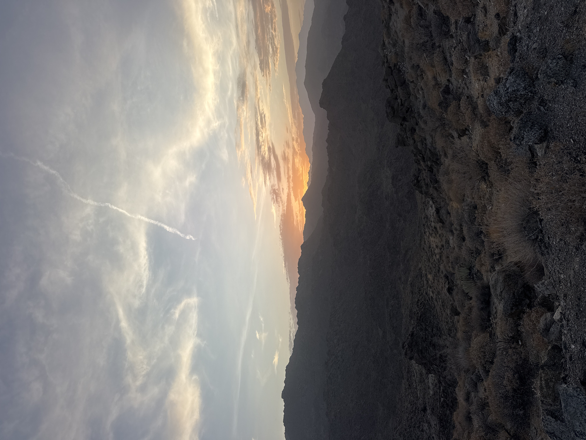 The image depicts a desolate, rocky desert landscape under a vast sky at sunset. In the foreground, dry, scrubby vegetation and dark, jagged rocks dominate the terrain. Beyond this, a series of dark, silhouetted mountain ranges recede into the distance. The sky is a canvas of soft colors, transitioning from pale blue and white at the top to warm oranges and pinks near the horizon, where the sun is setting behind the mountains. A faint contrail stretches across the upper portion of the sky. The overall impression is one of a remote and rugged natural environment. The location is not specified within the image itself, and there is no indication of the city of New Santa Rosa, United States of America. There are no people or specific actions visible in the scene.