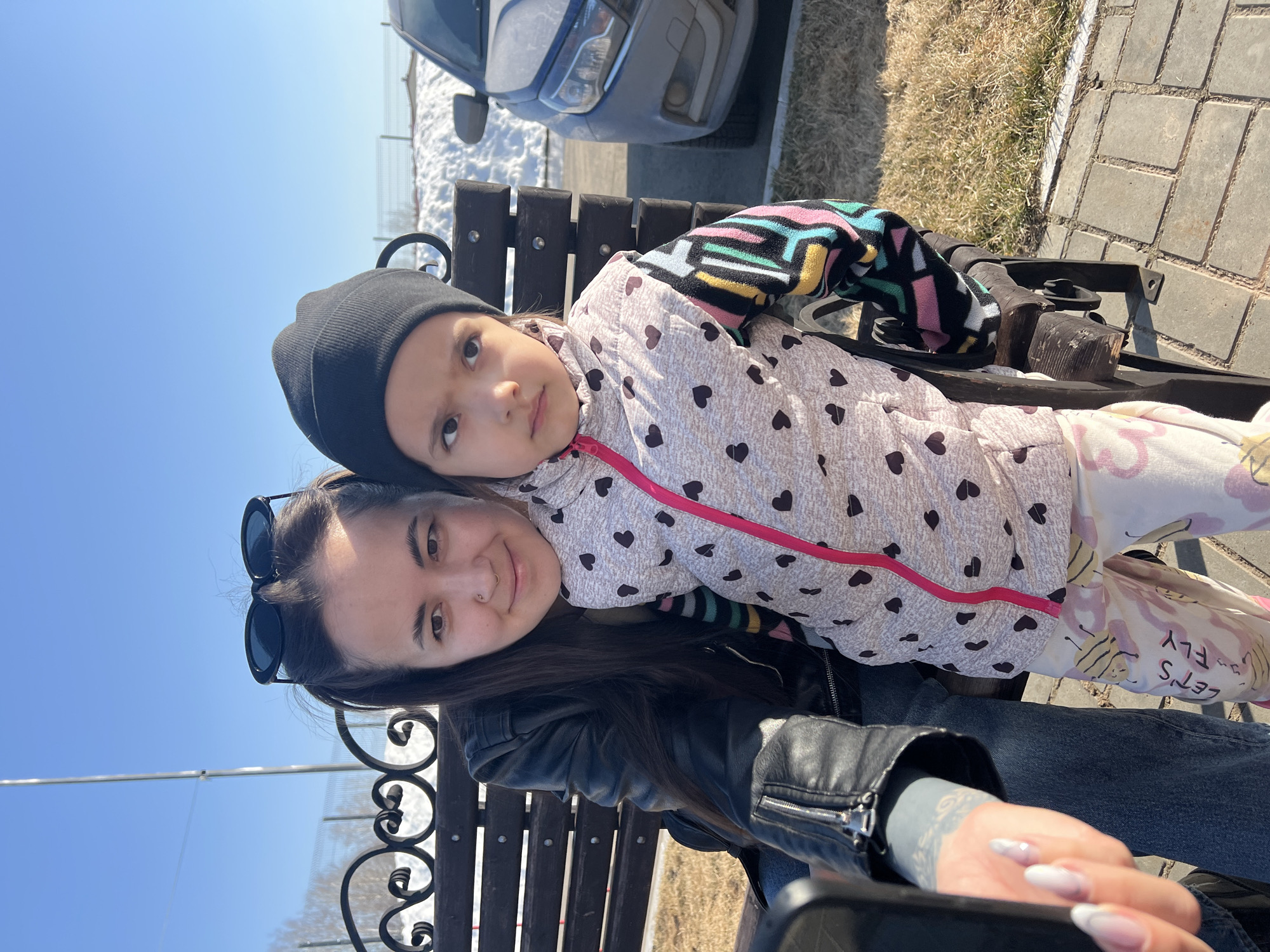 A woman and a young girl are sitting on a park bench. The woman has dark hair and is wearing a black leather jacket with sunglasses perched on her head. The girl is wearing a black beanie and a white jacket with black hearts and a pink zipper, with a colorful sleeved shirt visible beneath. They are close together on the bench. The background shows a car, a snowy area, a brick walkway, and a clear blue sky. The location may be Noviy, Russia.