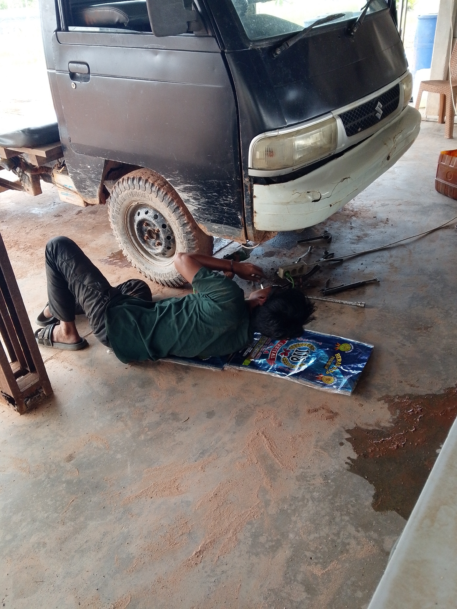Location: Bukit Ketok, Indonesia. A man lies on his back on a blue plastic sheet, apparently performing maintenance on the underside of a black motor vehicle. The vehicle has a muddy tire and a damaged white bumper. Tools are scattered on the floor nearby. The floor is concrete and covered with reddish-brown debris.