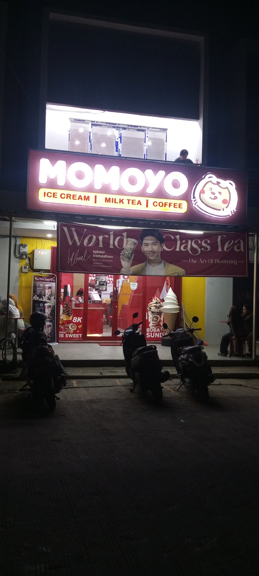 A nighttime exterior shot shows the front of a MOMOYO ice cream and milk tea shop in Setu, Indonesia. The prominent neon sign above the shop reads "MOMOYO" in large, bright pink letters, with smaller text below indicating "ICE CREAM | MILK TEA | COFFEE." To the right of the sign is a cartoon logo of a smiling bear character.
Below the main sign, a large maroon banner features a picture of actor and musician Iqbal Ramadhan, who is identified as the MOMOYO Brand Ambassador. He is holding a drink cup and the banner reads "World Class Tea -The Art Of Blooming-".
The shop's entrance is a red-framed glass door, with a yellow wall section to the left and a greyish-white wall to the right. Inside the shop, various promotional posters and menu boards are visible, along with a visible ice cream display and a person working behind the counter. Two large ice cream cones are displayed prominently outside the shop entrance, one with chocolate and cream swirls and another plain white one in a cone.
In front of the shop, on the paved sidewalk, are three parked motorcycles. The overall scene suggests a commercial establishment operating in the evening, likely attracting customers with its illuminated signage and displayed offerings.