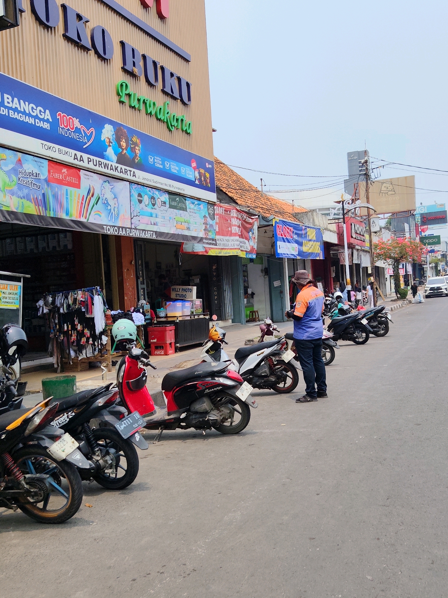 The image shows a street scene in Purwakarta, Indonesia, in front of a bookstore named "TOKO BUKU AA PURWAKARTA". The building facade has large, blue, raised lettering spelling out "TOKO BUKU" and green lettering spelling "Purwakarta". A blue banner with "100% INDONESIA" is displayed above a smaller banner advertising art supplies with images of coloring books and pencils.
Several motorcycles are parked along the side of the road. In the foreground, there are three motorcycles, one red, one black and white, and one black. Further down the street, more motorcycles are visible.
A man wearing a brown hat, an orange and blue shirt, and dark pants is standing on the street, looking at his phone. He appears to be the only person actively engaged with an object in the scene. Other individuals are visible in the background near the storefronts, some walking and others standing.
The street itself is paved with asphalt. The environment includes storefronts with awnings, utility poles with wires, and some vegetation. The sky is overcast and bright. The overall impression is that of a typical commercial street in a town or city.