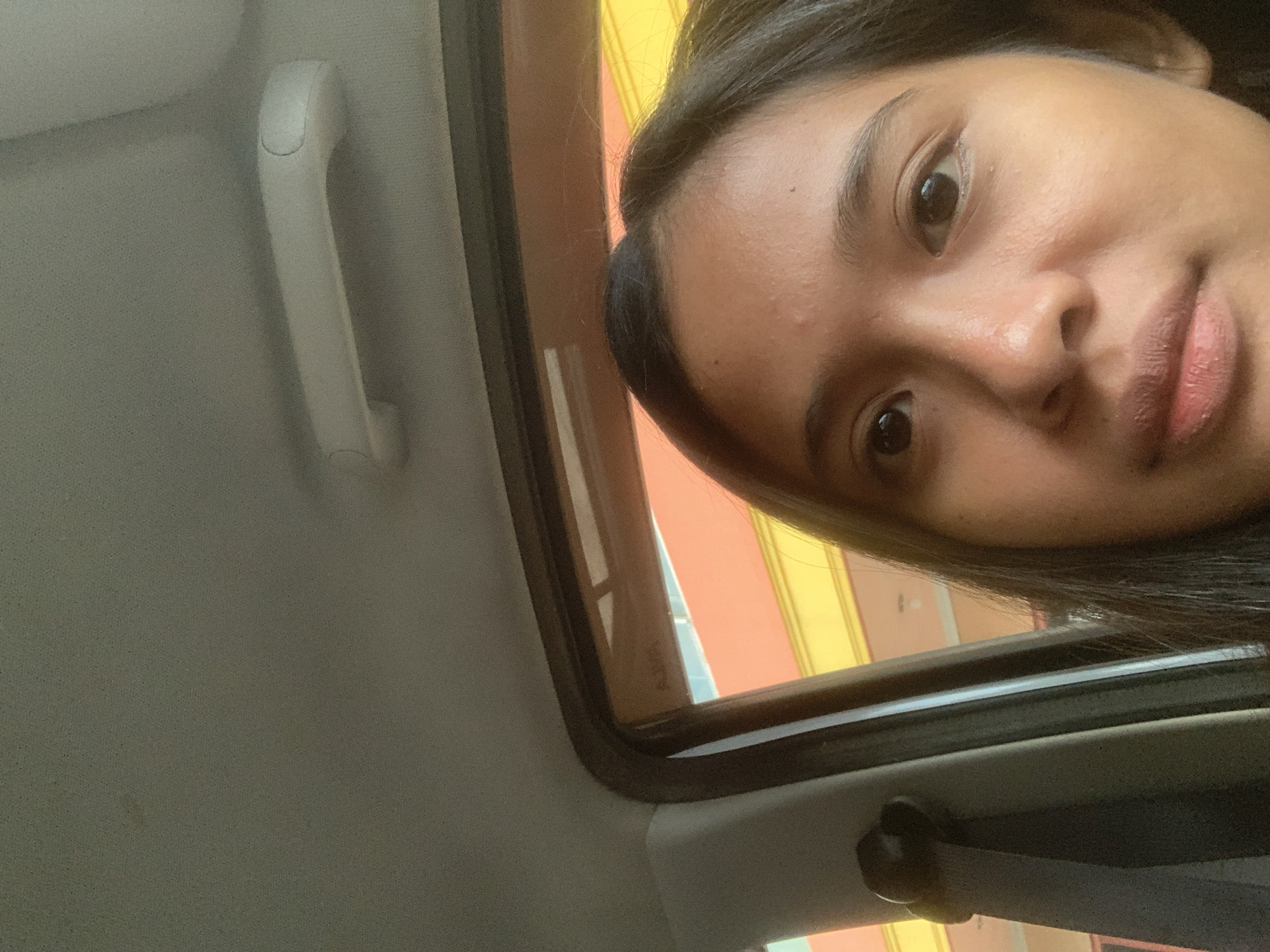 A selfie of a young woman with dark hair, taken inside a vehicle. Her face is positioned in the upper right quadrant of the image, with a portion of her dark hair visible. The background shows the interior of a car, including a grey ceiling, a white grip handle, and a window frame. Outside the window, stripes of orange and yellow suggest a building or structure, possibly in Samarinda, Indonesia.