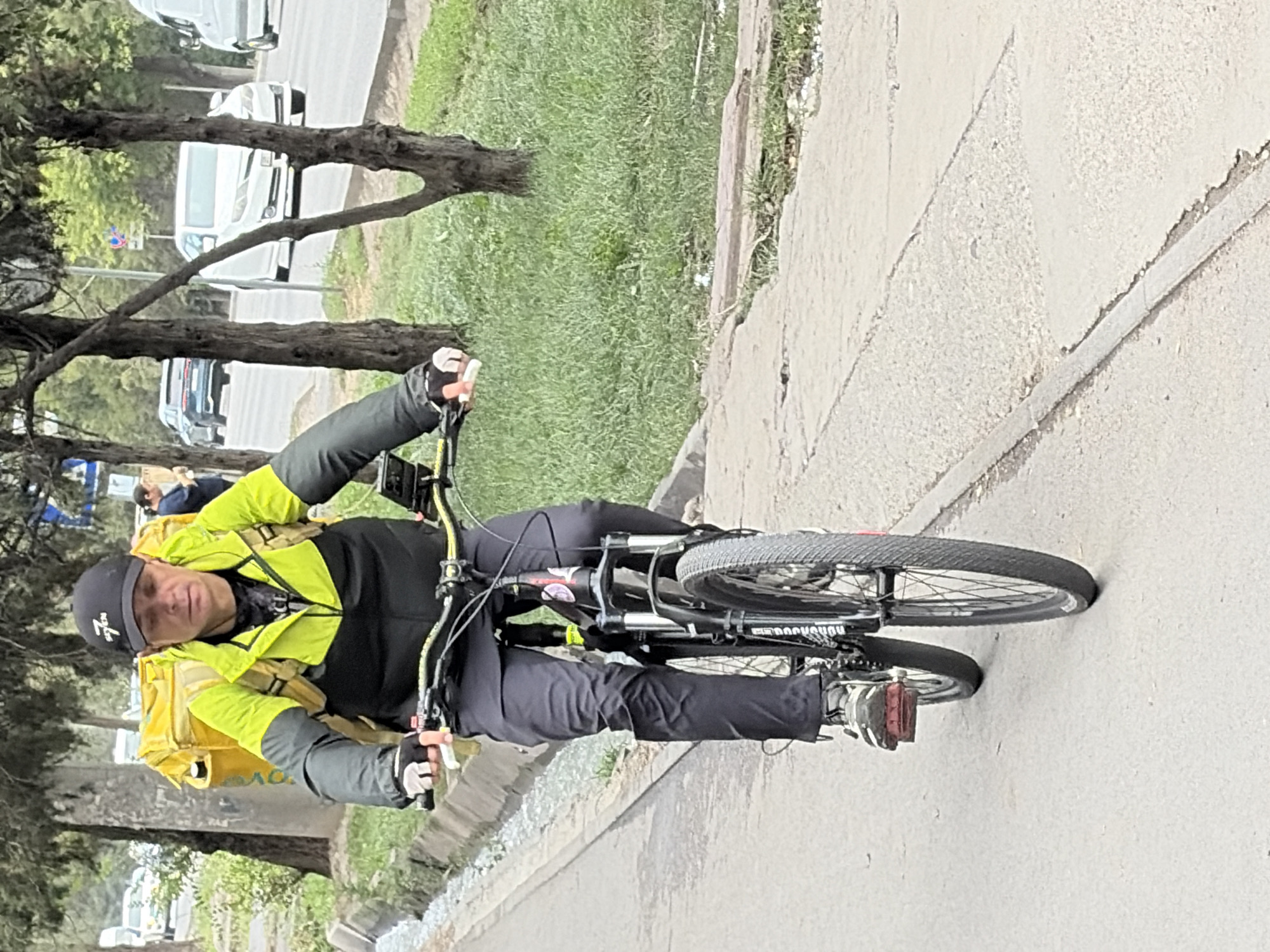 A man wearing a black baseball cap with a white logo, a lime green and black jacket, dark pants, and cycling gloves is riding a black mountain bike. He has a large yellow backpack strapped to his back. The man is on a paved path next to a street with cars driving on it. There is a grassy area with trees between the path and the street. The lighting suggests it is daytime.