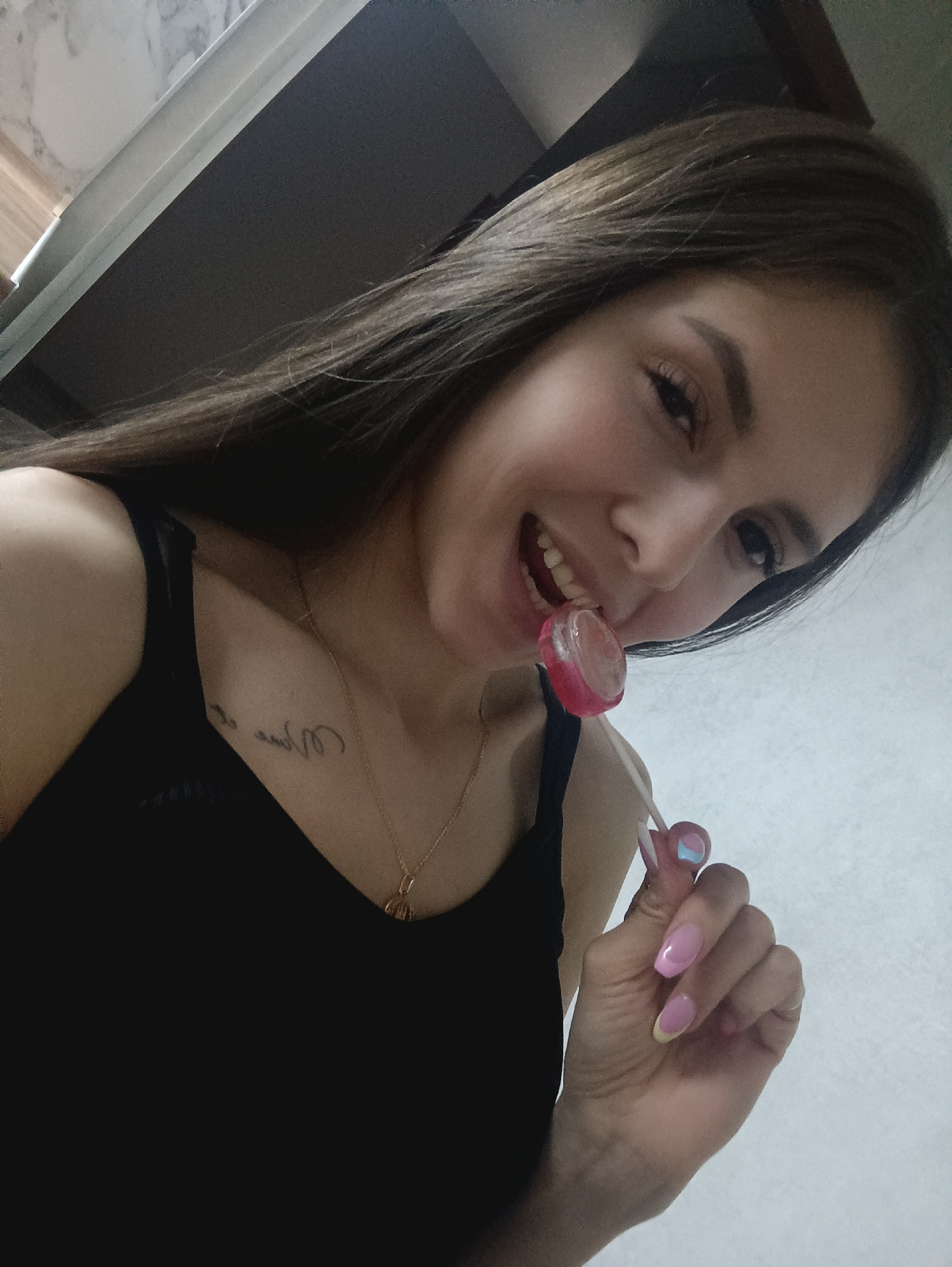 A young woman with long brown hair is biting into a red lollipop in an indoor setting. She is wearing a black tank top and a gold necklace. A tattoo is visible on her collarbone. Her fingernails are painted pink with a light blue accent on one finger. The background shows a light-colored wall and a glimpse of a dark grey surface above, possibly a cabinet or shelf. The location is in the city of Unknown, Unknown.