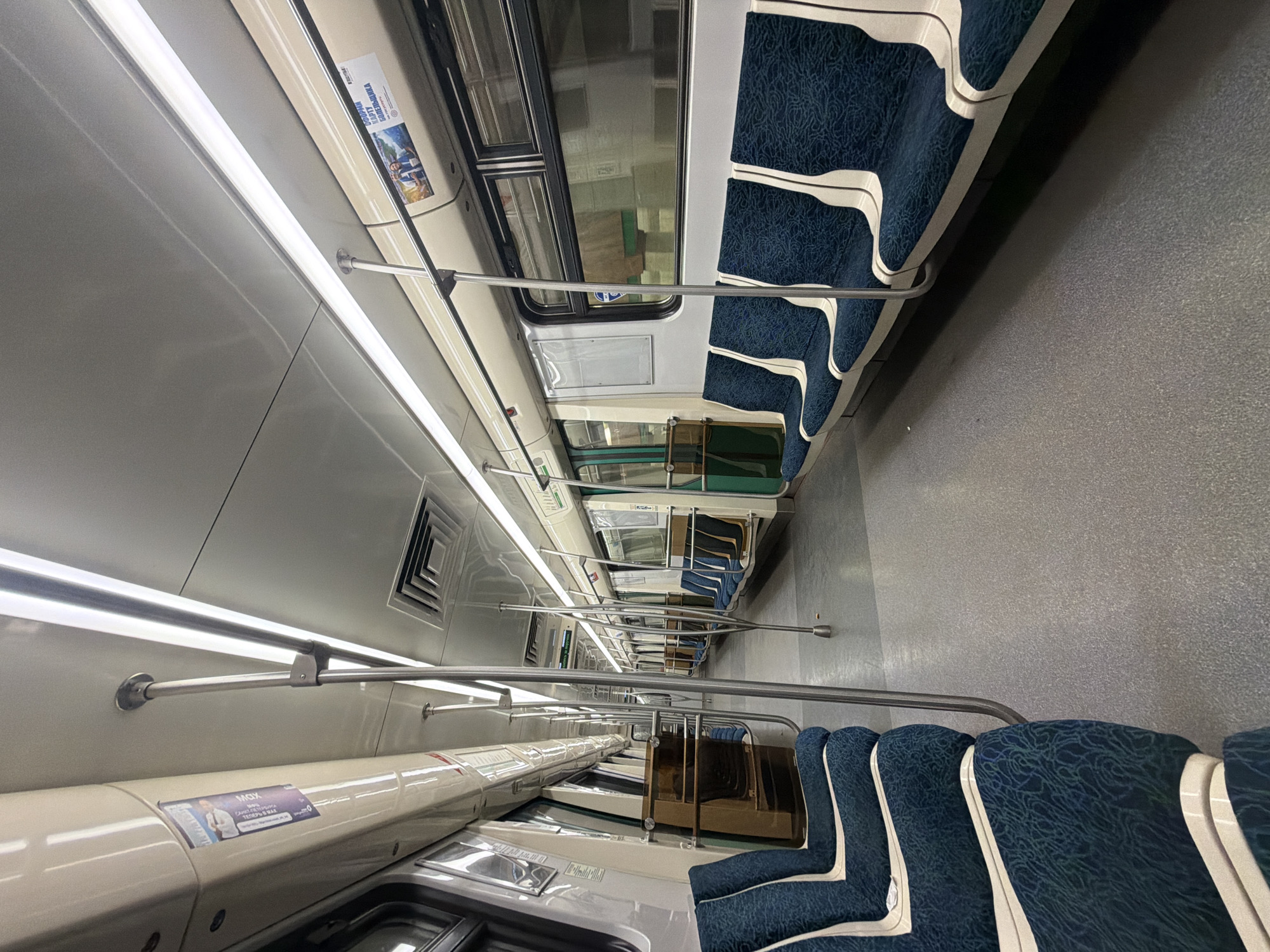 The image shows the interior of a metro train car, likely in Saint Petersburg, Russia. The car is mostly empty, with rows of seats upholstered in a blue patterned fabric and white trim on both sides. Handrails run along the length of the car for standing passengers. There are advertisements on the walls. The floor is a grey color. The lighting is provided by fluorescent lights that run the length of the car's ceiling. The train appears to be in motion based on the blurred background seen through the windows.