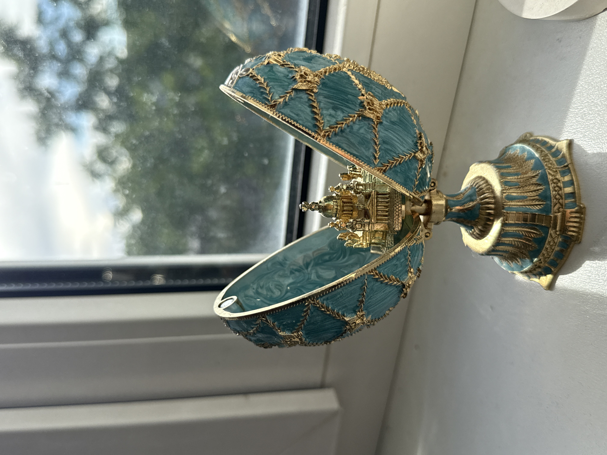 An ornate, open Fabergé-style egg, likely made of gilded metal and enamel, rests on a white surface. The egg is decorated with intricate gold patterns against a teal background. Inside the egg, a miniature golden palace or cathedral complex is visible. The egg sits on a matching gilded stand. The background features a window with vertical blinds and a blurred view of green trees and the sky, suggesting an indoor setting near a window. The lighting indicates it is daytime. There is no indication of this scene being in Volgograd, Russia.