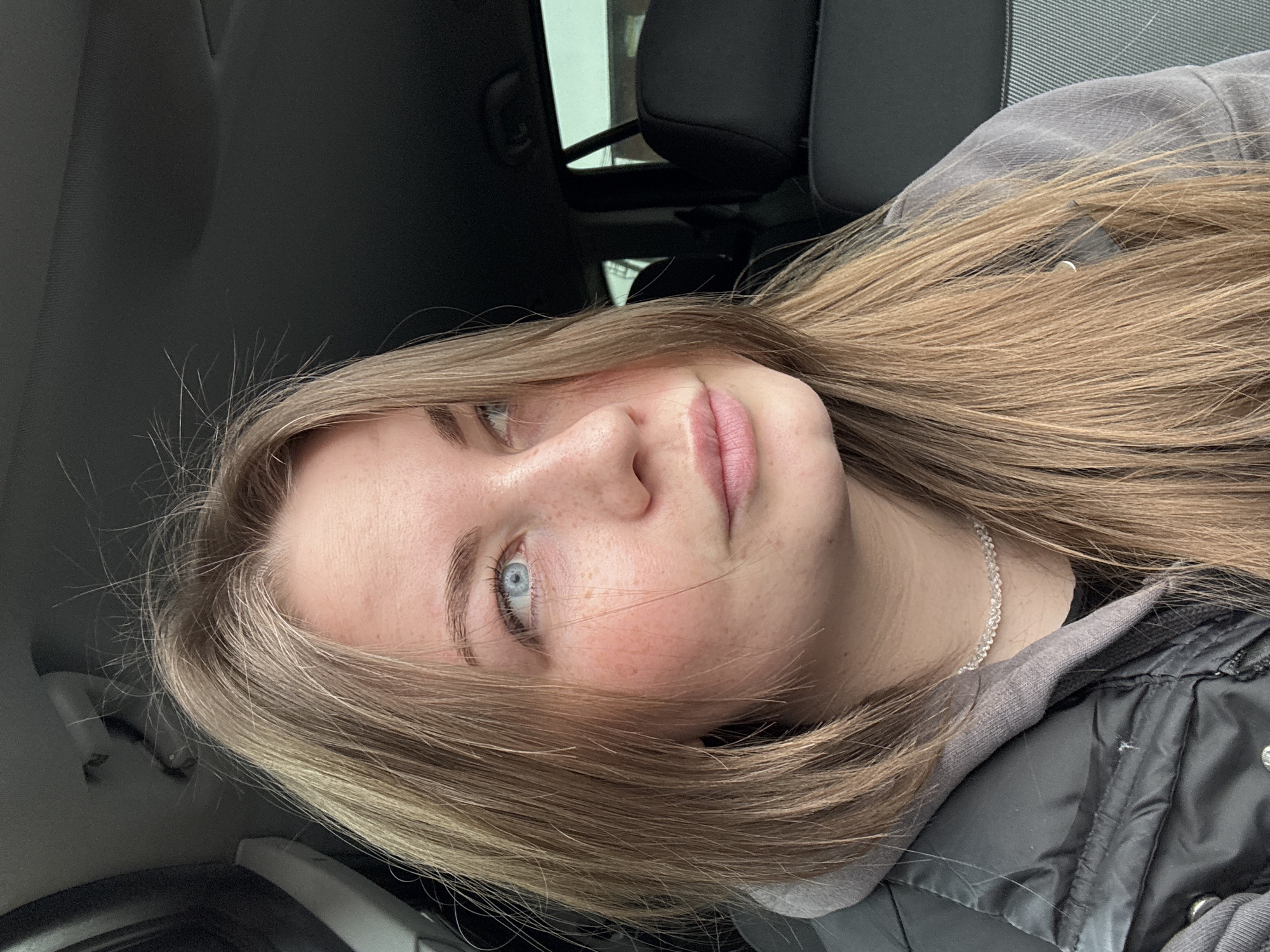 A young woman with blonde hair and blue eyes is looking at the camera. She is wearing a grey hoodie and a black vest. She is sitting in a car. The location is unknown, but it is not in Фоки, Russia.