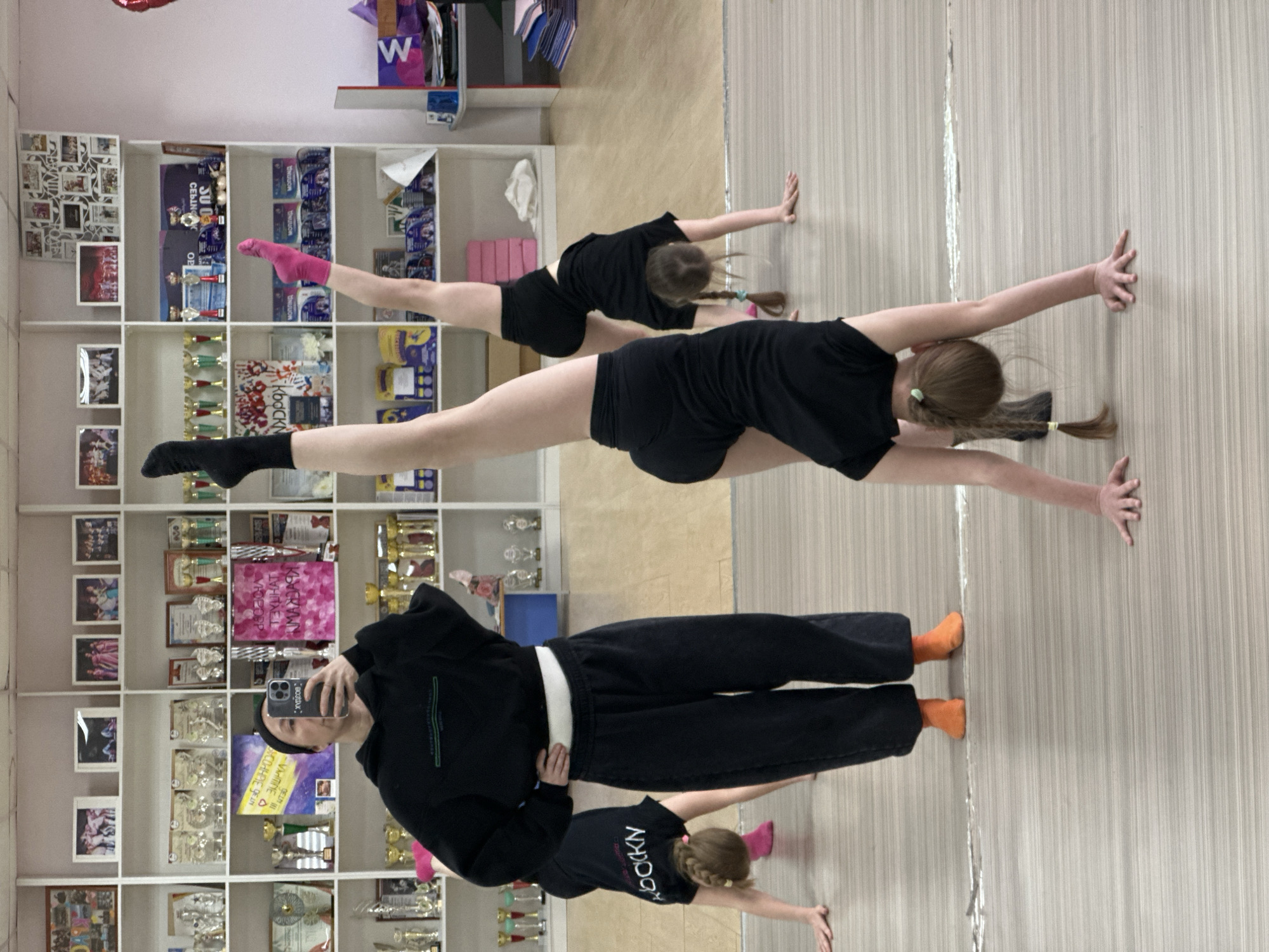 The image depicts an individual taking a selfie in a room, likely a dance or gymnastics studio, as evidenced by the presence of two young individuals performing splits in the background. The room appears to be located in Votkinsk, Russia, indicated by the text "bOCKN KLUCKYWN", possibly a dance school or studio with that name, as well as other text written in Russian. Shelves filled with trophies, awards, and various decorative items are visible behind the person taking the selfie. The overall setting is one of a practice space or studio with achievements showcased on the shelves. The selfie taker is wearing black clothes and is positioned in front of the shelves, while the young performers are on the floor near a striped wall.
