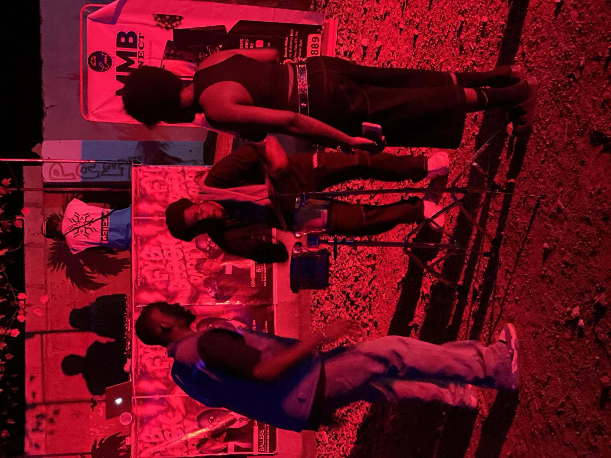 The image captures an outdoor event at night, bathed in vibrant red lighting. In the foreground, three individuals are visible. A person on the left, wearing a blue vest over a dark shirt and light-colored pants, is in motion, with their back to the camera. To their right, a person in dark clothing, including a black t-shirt and black pants, stands facing away from the viewer, looking towards the left. On the far right, a third person, also in dark clothing, wears a black tank top and dark pants, with their back turned to the camera.
Behind the individuals, a DJ setup is visible, featuring a laptop and DJ equipment. Posters with images of people are displayed on a backdrop. A banner with the text "MMB CONNECT" and a logo is prominent on the right side of the image. The ground appears to be covered in gravel or small stones. The overall scene suggests a lively outdoor gathering or performance. The location is identified as Durumi, Nigeria.