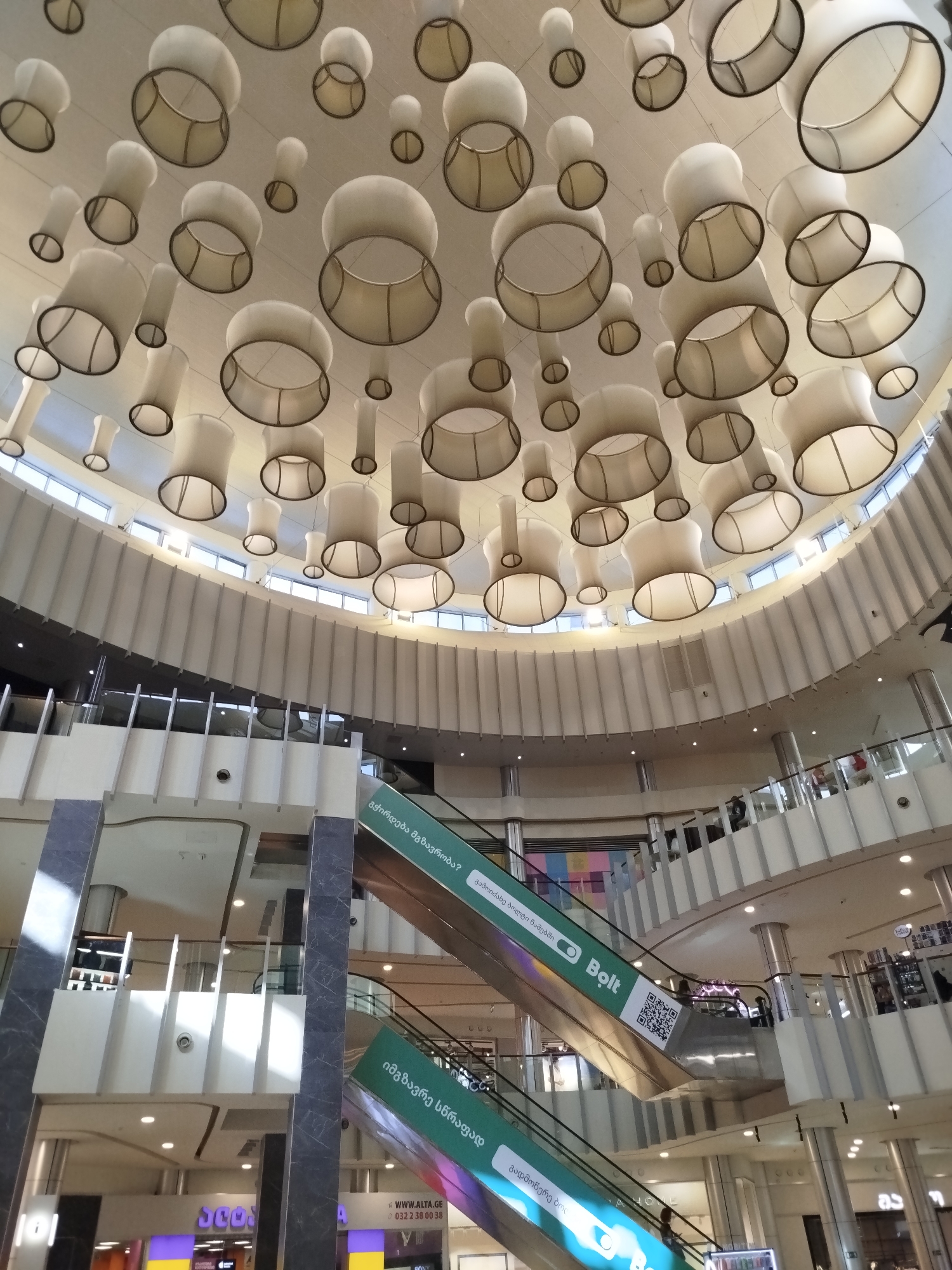 This image captures the interior of a modern shopping mall in Tbilisi, Georgia. The focal point is the impressive ceiling, adorned with numerous large, white, drum-shaped lamps suspended at varying heights. These lamps, with their dark rim detailing, create a dramatic and visually striking effect.
Below the ceiling, the mall's multi-level structure is evident, with escalators and elevated walkways visible. People are seen moving about on different levels, indicating a typical day with shoppers engaged in their activities. The architecture features clean lines, with white and gray elements dominating the interior design, accented by polished metal columns.
Several signs are visible, including green escalators with text in Georgian and English. One sign prominently displays the "Bolt" logo and a QR code, suggesting the availability of ride-sharing services. Another sign directs to "WWW.ALTA.GE", a Georgian electronics retailer. The presence of "SONY" branding further hints at the types of stores found within the mall. The lighting is bright and diffuse, typical of an indoor commercial space. The overall impression is one of a bustling, contemporary shopping environment.