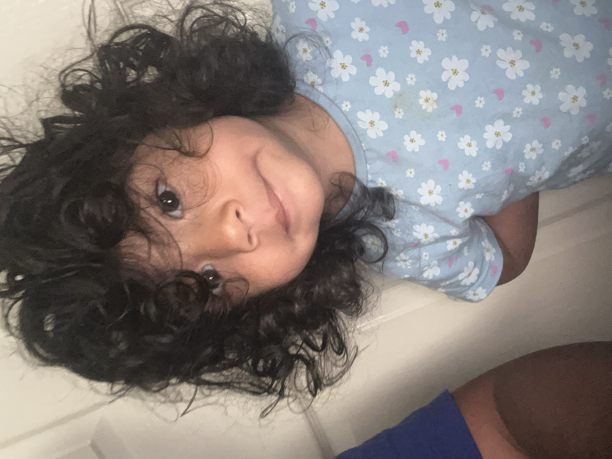 A close-up, slightly angled indoor shot shows a young child with curly dark hair and brown eyes. The child is wearing a light blue t-shirt with a pattern of white flowers and small pink hearts. A portion of a blue garment is visible on the left side of the frame, and the child's legs are partially visible. The background consists of a light-colored wall. The image does not contain any elements that would allow for the determination of its location within Boston, United Kingdom, or any other city.