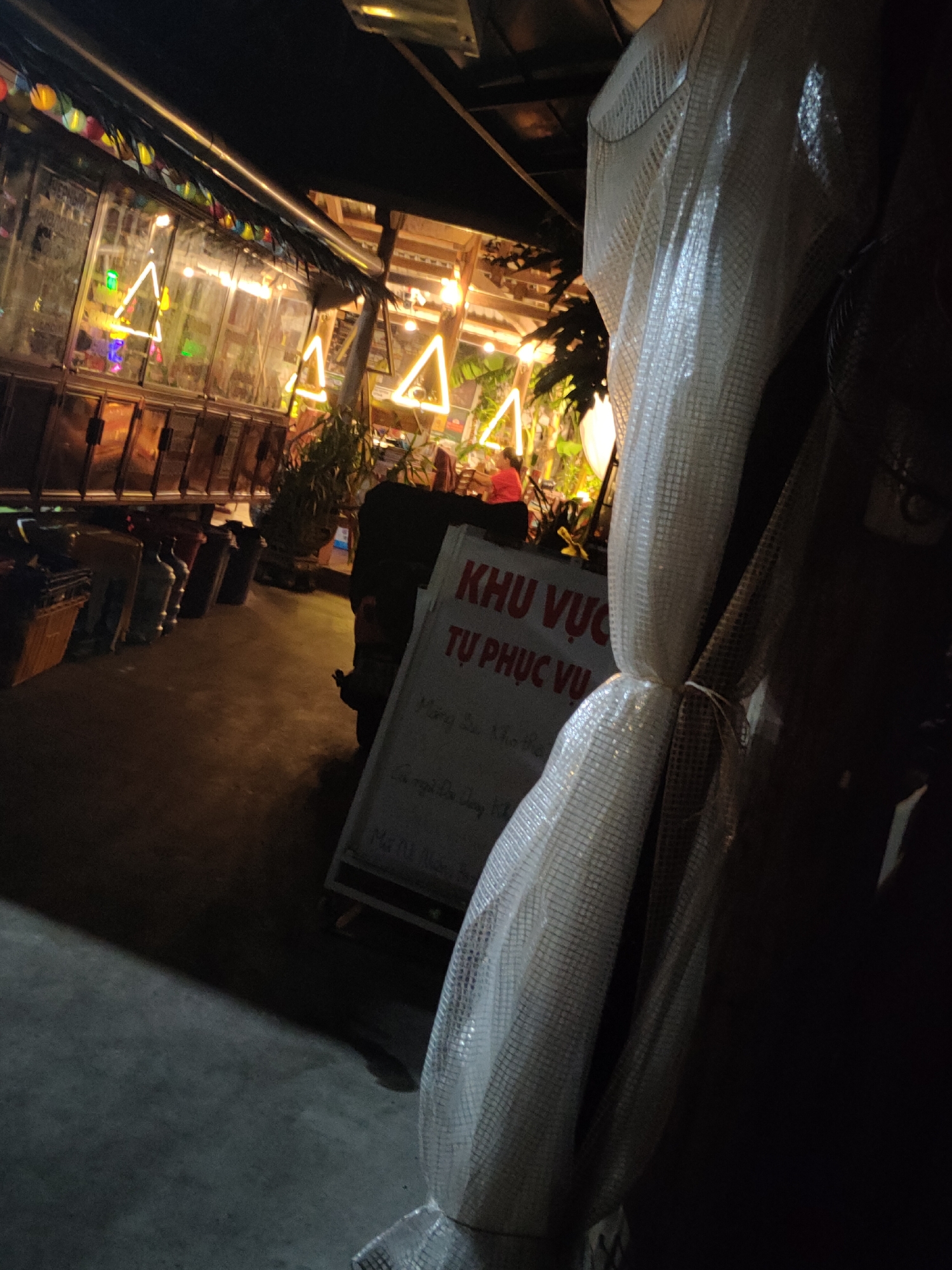 The image depicts an outdoor scene at night, likely a market or a food stall area, with a sign that reads "KHU VUC TU PHUC VU". There are illuminated triangular neon signs in the background, as well as strings of colorful lights. In the foreground, a white, netted material is draped, obscuring part of the scene. A white board with Vietnamese text is visible on the right side of the image. The ground appears to be concrete. The overall ambiance suggests a lively evening setting.