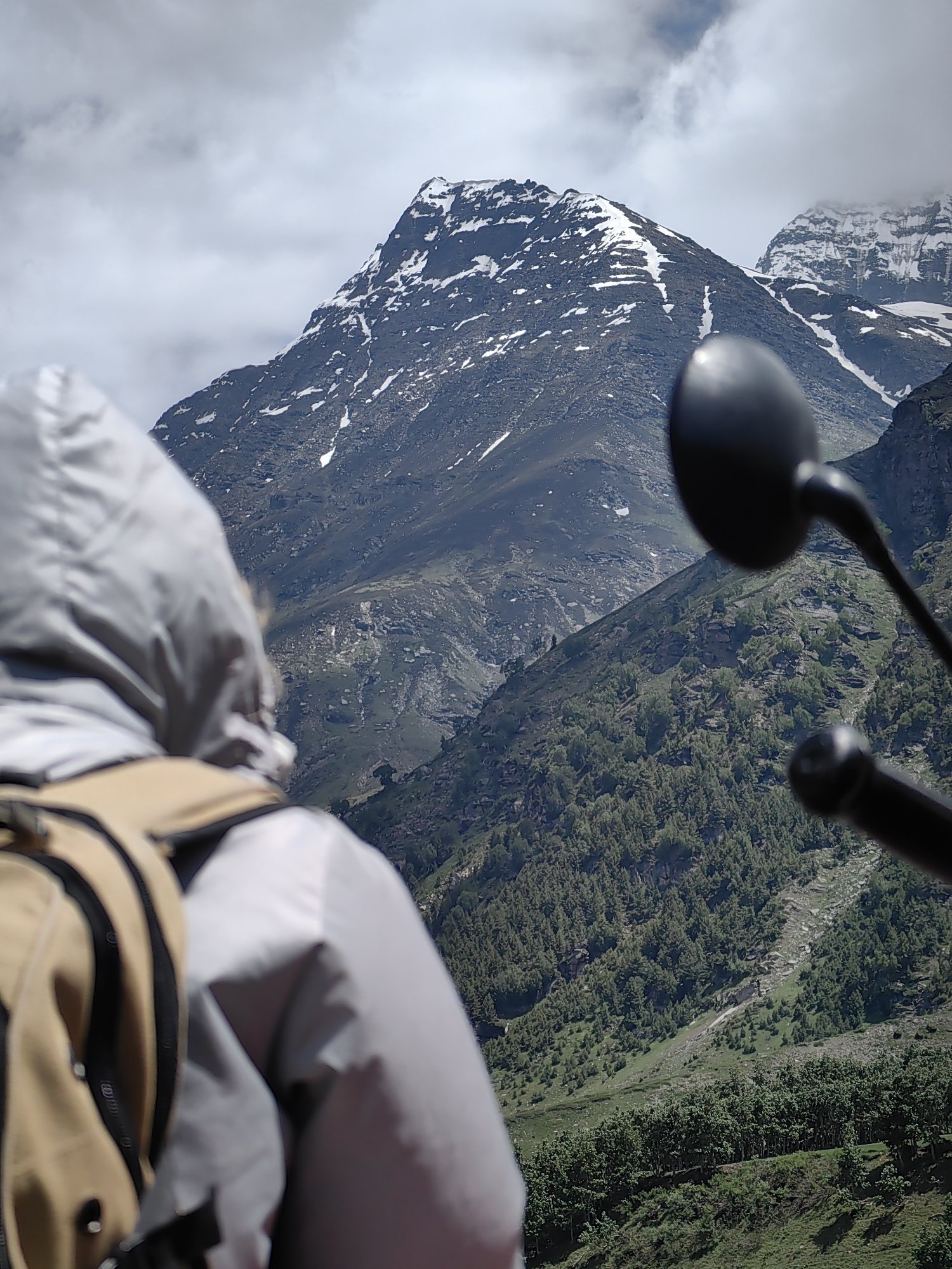 A person wearing a grey hooded jacket and a beige backpack is positioned in the foreground, obscuring the lower portion of a majestic mountain range. The mountains, partially covered in snow, dominate the background, with lush green vegetation clinging to their slopes. The sky is partly cloudy, with grey clouds gathering. In the immediate foreground, a black motorcycle rearview mirror is visible on the right side of the frame. The image appears to have been taken from the perspective of a rider on a motorcycle, looking towards the mountainous landscape. The location is identified as Burwa, India.