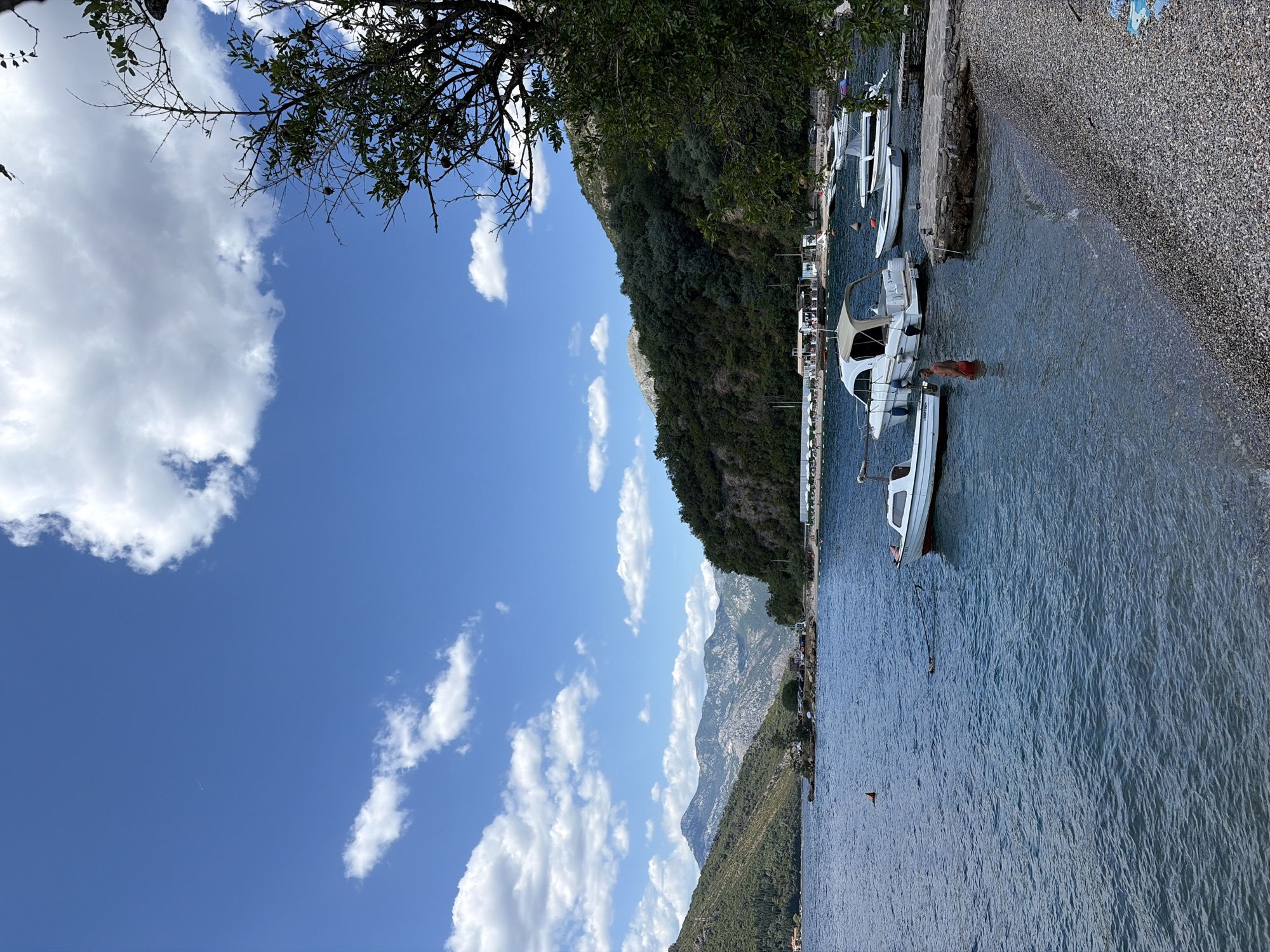 The image depicts a scenic view of the Bay of Kotor in Lepetane, Montenegro, on a clear day with scattered white clouds against a bright blue sky. In the foreground, a pebble beach transitions into the calm, blue water. Several boats are moored or anchored in the bay. A white motorboat with a canvas canopy is prominently positioned, and a smaller white boat is closer to the shore. A person, wearing red shorts, is standing in the water near the smaller boat, partially submerged. Further out in the bay, more boats are visible, and a small island with buildings and parked cars can be seen in the distance. The background is dominated by lush green, tree-covered mountains rising from the water's edge, with jagged, rocky peaks visible in the far distance. A tree with branches and green leaves partially frames the right side of the image.