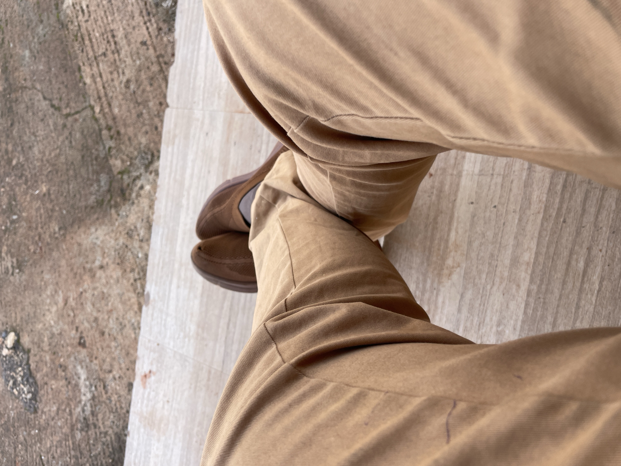 The image shows a close-up, high-angle shot of a person's legs wearing tan trousers and brown shoes, resting on a light-colored tiled floor. The background is blurred, showing a rough, reddish-brown concrete surface with some scattered pebbles and a small red stain. The lighting appears to be natural daylight. The overall scene is simple and focused on the lower body and footwear. There is no visible indication of the specific location in Jos, Nigeria.
