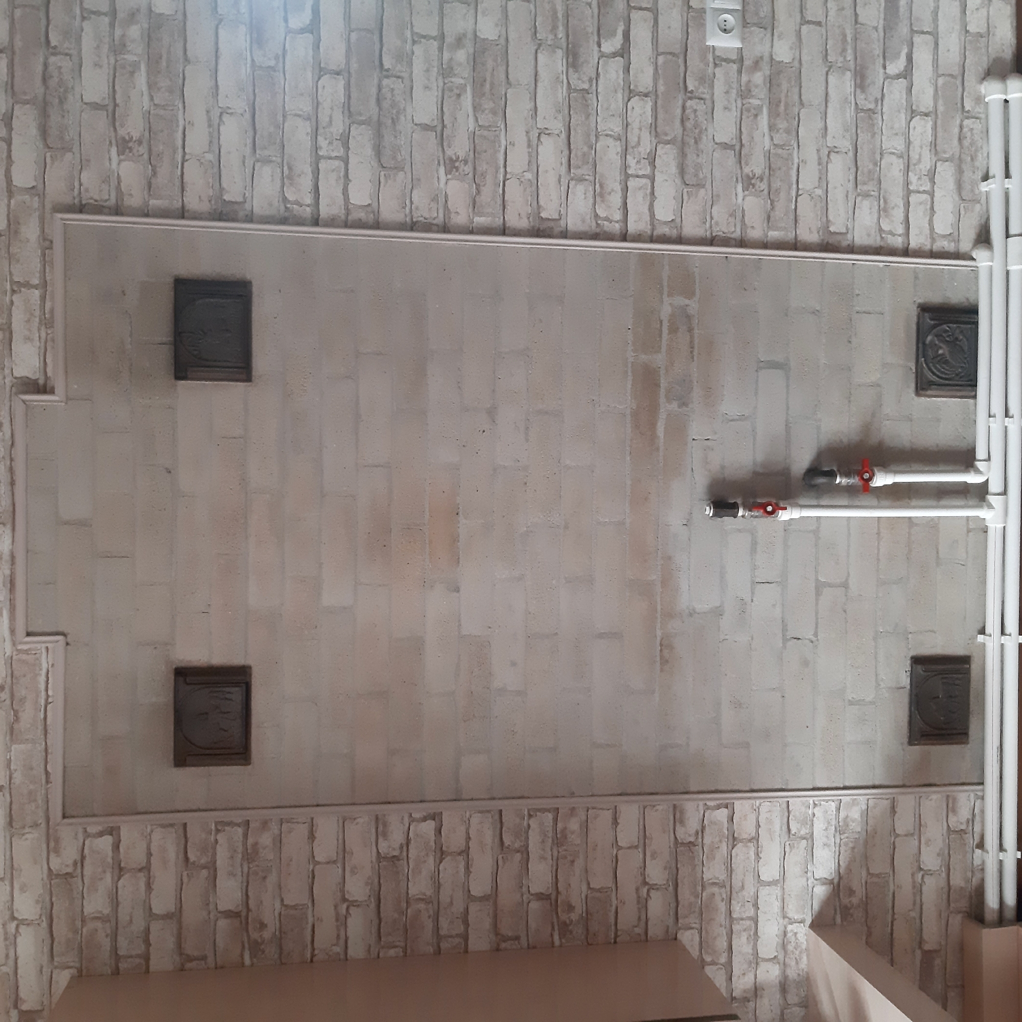 The image captures an interior view, looking upwards at what appears to be a ceiling or high wall. The surface is predominantly covered in light-colored, faux brick or brick-patterned wallpaper/paneling.
A large, lighter rectangular section is framed by simple, light-colored molding, standing out against the brick pattern. Within this framed area, four dark, square-shaped decorative plaques are symmetrically mounted. Each plaque features an embossed circular design, possibly depicting a stylized face or creature, though the details are not perfectly clear. Two plaques are on the left side and two on the right.
To the right of the central framed section, a system of white utility pipes runs vertically before branching into two horizontal segments. On these horizontal pipes, two red-handled ball valves are visible, along with a black fitting, possibly a pressure gauge or another type of valve. Above this pipe work, on the brick-patterned wall, a white electrical outlet with two circular sockets is present.
At the bottom of the image, parts of a light-colored, possibly wooden, structure or piece of furniture are partially visible, suggesting a surface or railing below the main wall. The overall lighting is bright, with some glare noticeable at the top. The scene depicts a functional indoor space, possibly in Yeghegnadzor, Armenia, blending rustic brick-like aesthetics with exposed utilities and decorative accents. No specific activities are taking place, and no discernible text is visible.