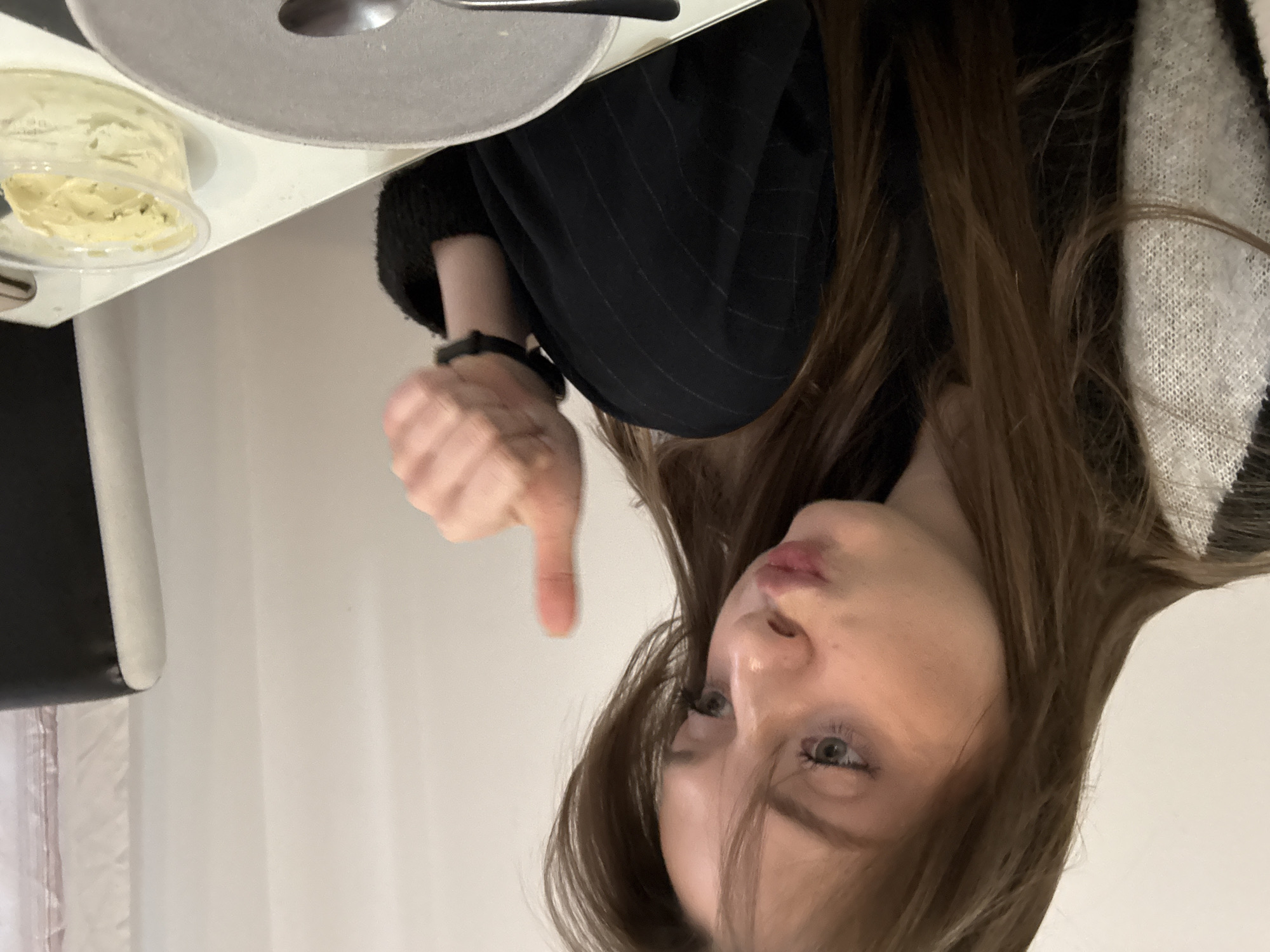 A woman is positioned behind a table, giving a thumbs-down gesture. A plate and a small container of food are on the table to the left. The background consists of a white wall and the edge of a chair. The location is not specified, so I cannot confirm it is in Kragujevac, Serbia.