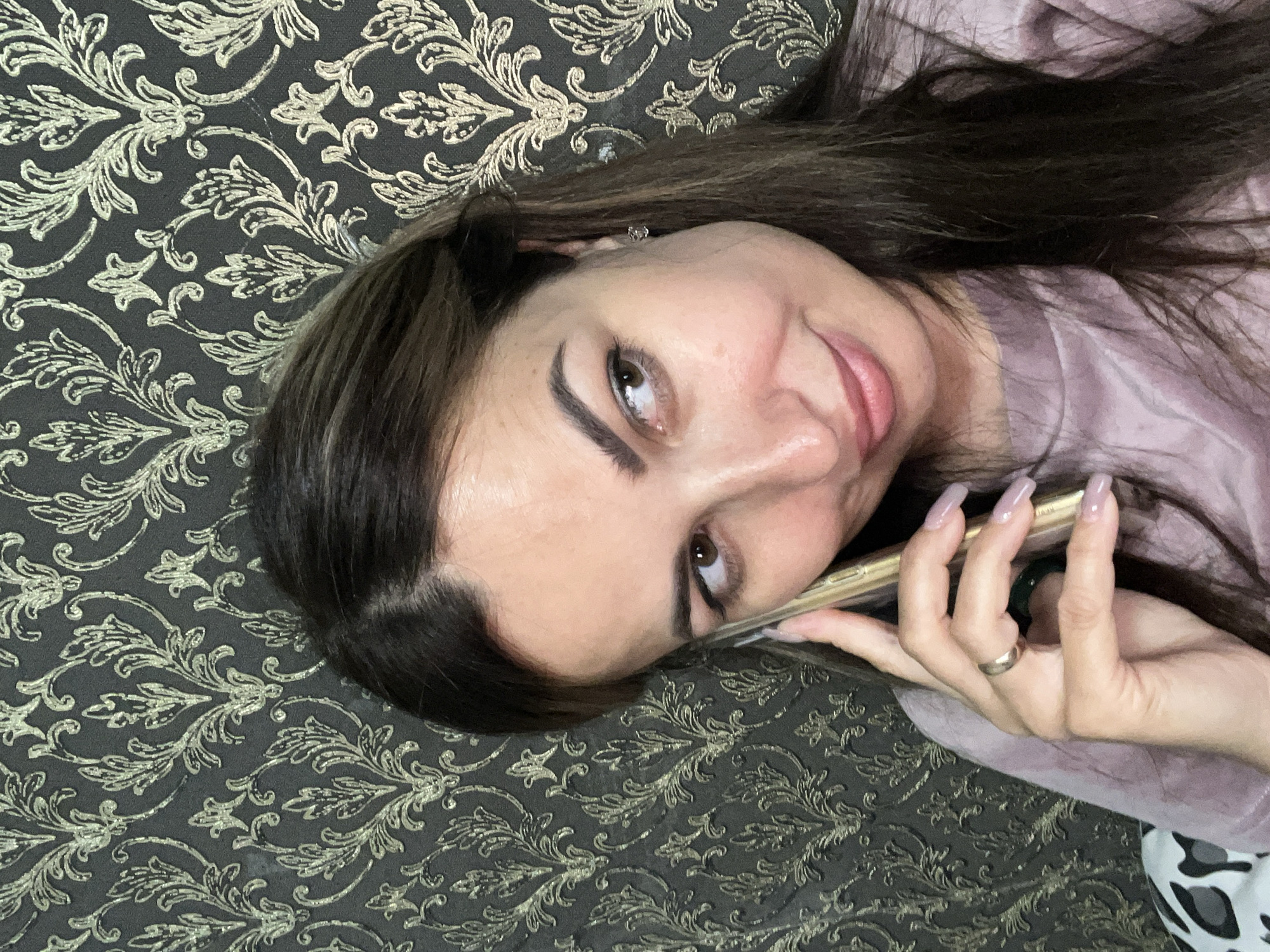 A medium shot of a woman with long, dark brown hair is taken from the chest up, as she holds a smartphone to her right ear and looks to her right. The woman is wearing a mauve colored shirt and has a ring on her left ring finger. The background is a dark wallpaper with a repeating gold floral pattern. The lighting is soft and the scene is indoors.