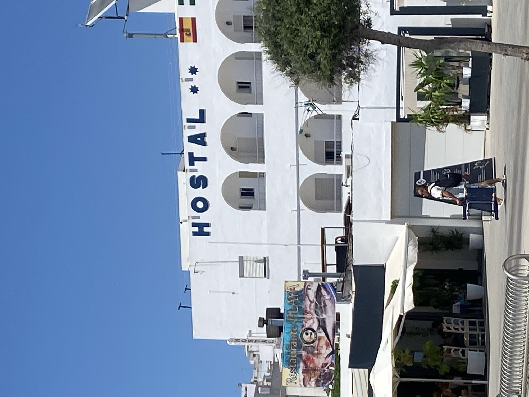 A clear, sunny day in Conil de la Frontera, Spain, showcases a white building with arched balconies and dark blue lettering spelling "HOSTAL" above the second-floor windows. Two star-shaped decorations are also present on the building. A Spanish flag and a green flag are mounted on the right side of the building. To the left, a sign for "Restaurante PLAYA" displays images of fish. A tree with dense green foliage grows in the foreground, partially obscuring the building. In front of the building, a person walks with a rolling suitcase and a backpack. A white awning covers an outdoor seating area to the left, where several people are seated at tables. A metal rack is visible in the lower left corner.