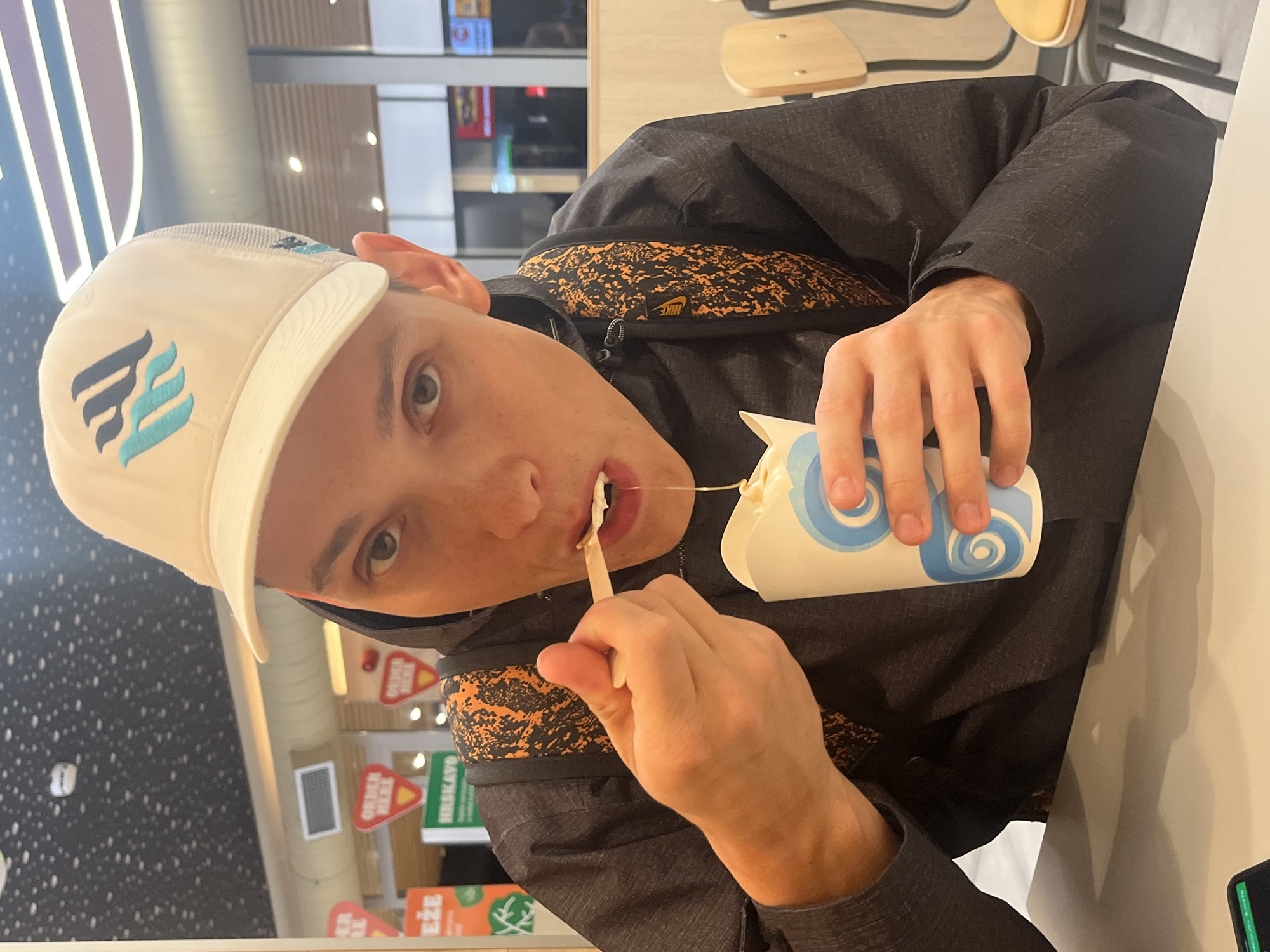 A person wearing a white baseball cap with a blue logo and a dark jacket with orange accents is eating a dessert from a white and blue container with a wooden spoon. The person is looking directly at the camera. The scene appears to be indoors, possibly in a restaurant or food court. There are signs and other people visible in the background, suggesting a public place. The location is in Budva, Montenegro.