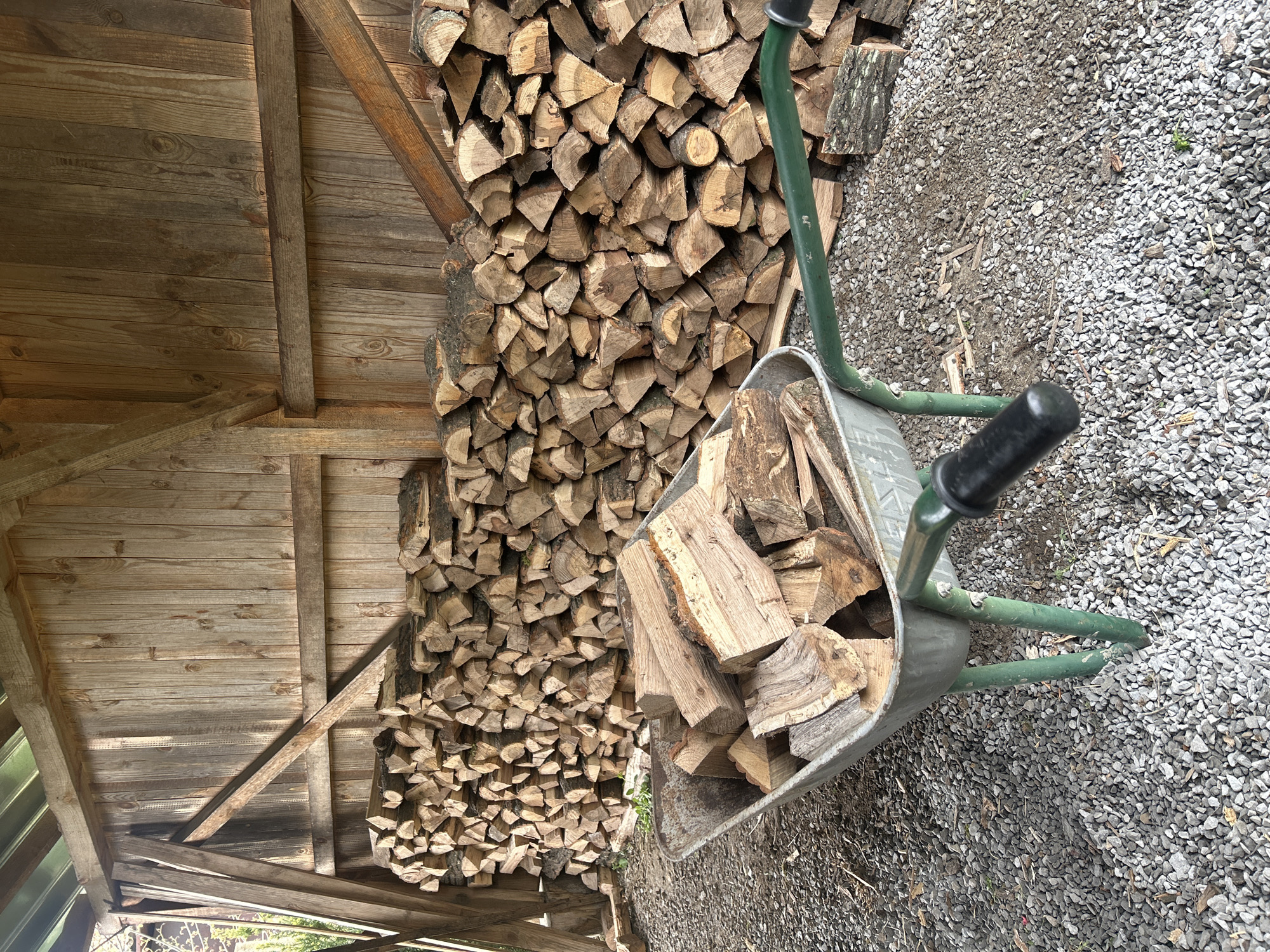 The image depicts a wood storage area, possibly in Senovo, Bulgaria. A large stack of split firewood is neatly arranged inside a wooden structure with a roof. A wheelbarrow, partially filled with more split wood, sits on a gravel surface next to the woodpile. The environment appears rural or semi-rural. No people are visible.