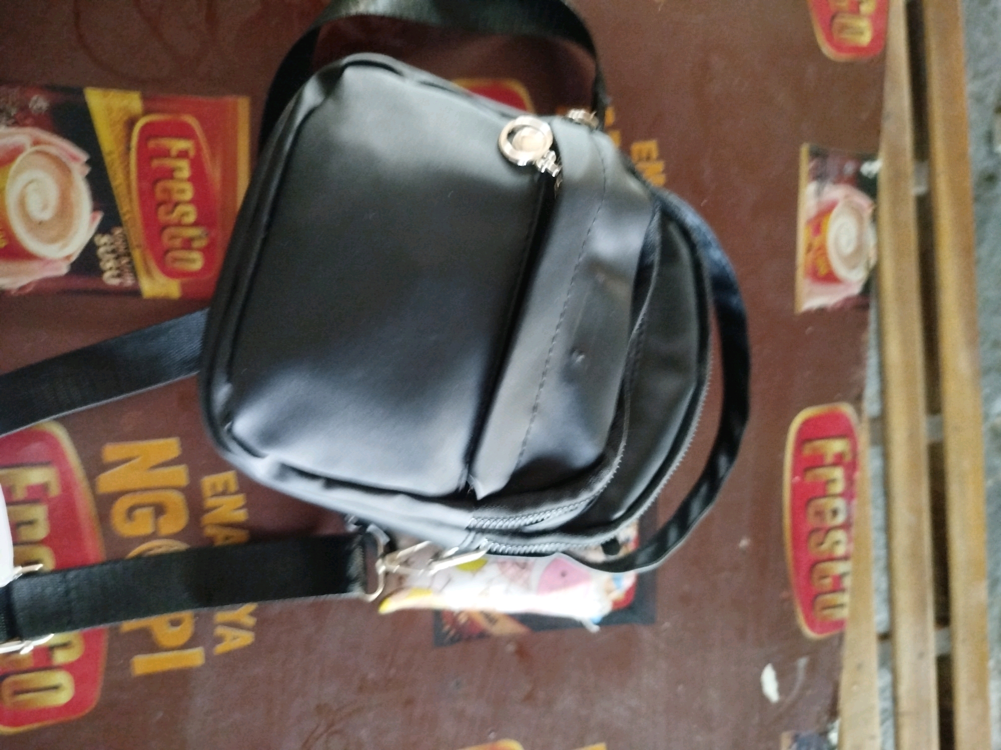A black sling bag is positioned diagonally on a brown surface that appears to be a table. The surface is decorated with what looks like packaging for a product called "Fresco" and other text that is partially visible and appears to be in Indonesian, possibly indicating "Ena Pi Ya Ngo" and "Gogo". There are also some colorful wrappers or stickers visible underneath the bag. To the right of the table, vertical wooden planks are visible, suggesting an outdoor or partially enclosed environment. The image does not provide enough information to determine a specific location within Gilang, Indonesia, beyond the general context of being on a table with Indonesian packaging. There are no people or animals in the image.