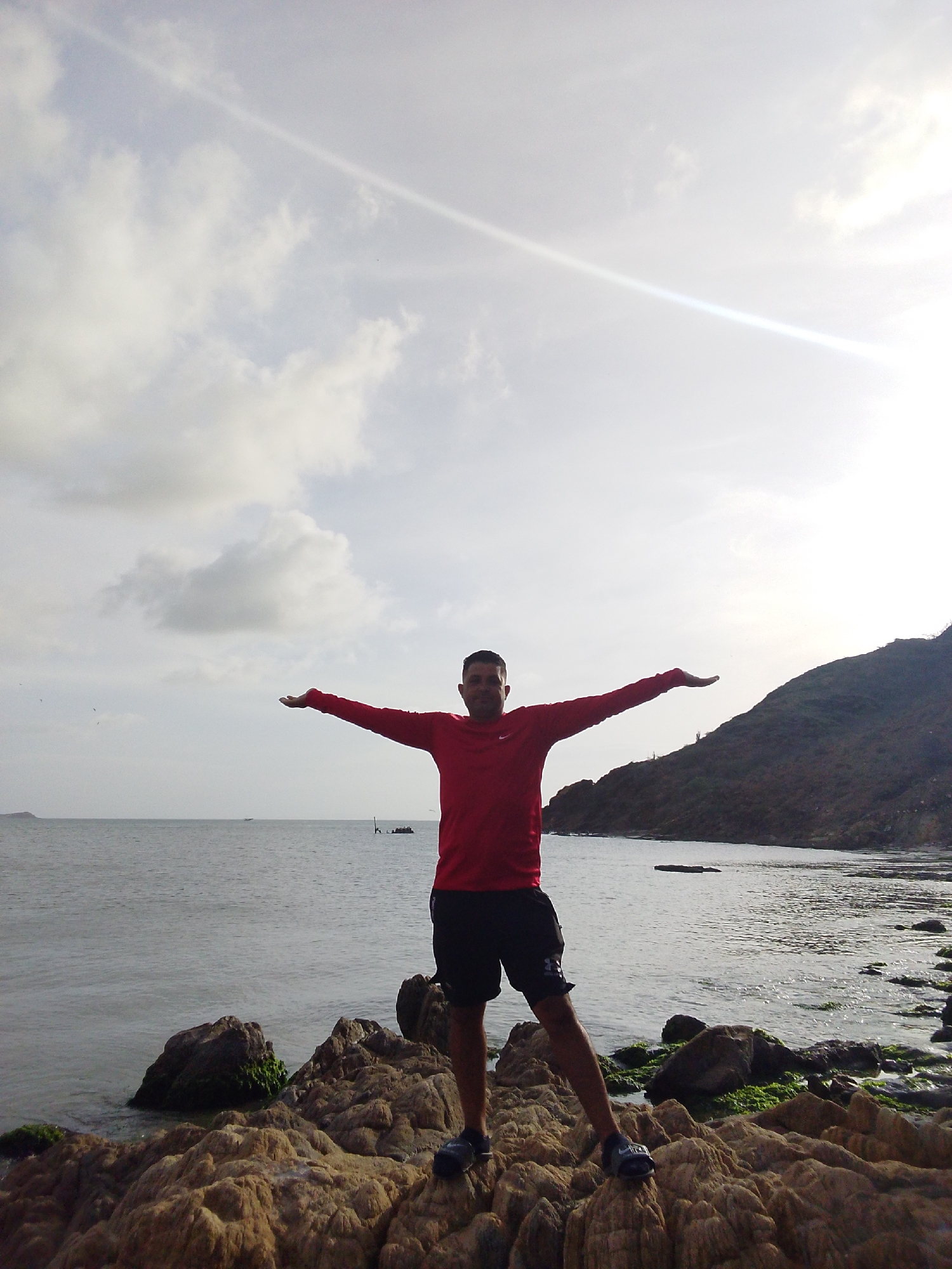 A full-body, medium shot captures a man standing on a rocky shore in Juan Griego, Venezuela, with his arms outstretched to his sides. The man is wearing a red long-sleeved shirt and black shorts. He is facing forward, towards the camera, with a slight smile. The background consists of the calm ocean and a lush, green hillside. The sky is partly cloudy with bright sunlight filtering through. In the distance, a small boat can be seen on the water. The foreground is dominated by the textured, uneven rocks of the shoreline, some covered with moss. The overall scene conveys a sense of freedom and connection with nature.