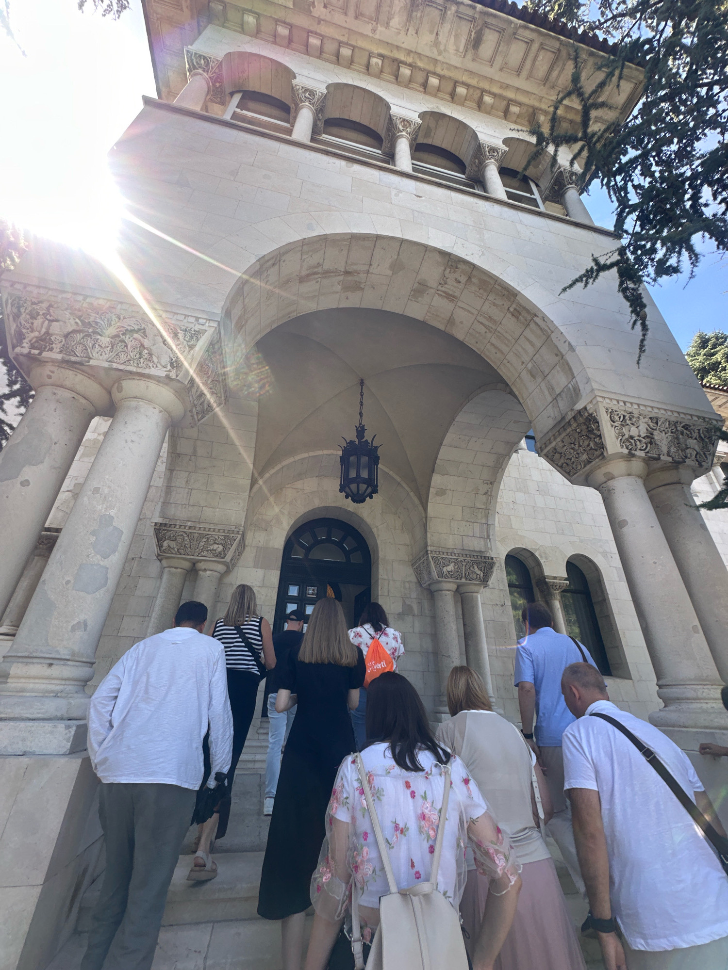This image captures a group of people ascending a grand stone staircase towards the entrance of a building. The architecture is characterized by large, fluted columns supporting an arched entryway with ornate carvings above. Sunlight streams in from the left, creating lens flare and highlighting the textured stone. A vintage-style lantern hangs within the archway.
The people are dressed casually, suggesting a daytime outing. A man in a white long-sleeved shirt and grey pants is leading the group, followed by a woman in a striped top, another woman in a black dress, and several others partially visible.
The building's facade appears to be constructed from light-colored stone blocks, with arched windows visible further back. The overall impression is one of historical significance and architectural grandeur.
This scene is located at the **National Museum of Serbia** in Belgrade, Serbia. The distinctive architecture, particularly the prominent columns and arches, is a hallmark of the museum's facade.