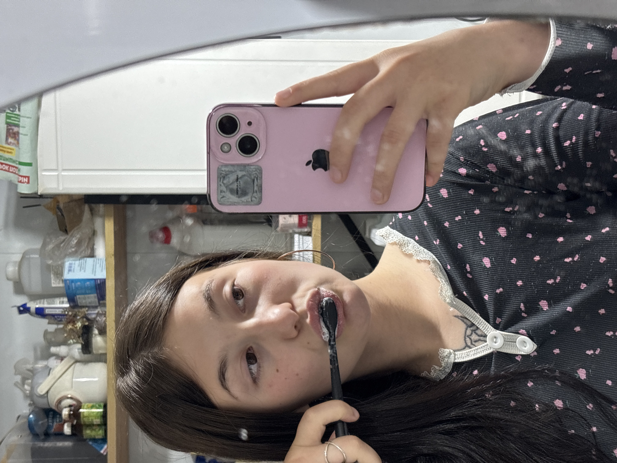 A young female is depicted in a mirror selfie, brushing her teeth, likely in a bathroom located in Фоки, Russia. She has long, dark hair and wears a dark-colored top featuring small pink floral or dot patterns and a white lace trim. A black toothbrush, laden with white foamy toothpaste, is positioned in her mouth, held by her left hand. A simple ring adorns the index finger of her left hand.
She uses her right hand to hold a pink iPhone, enclosed in a matching pink case, to capture the image in the mirror. The iPhone's dual camera lenses and the Apple logo are clearly visible on its back, along with a small, square, reflective sticker on the bottom left corner. The background, reflected in the mirror, reveals a cluttered shelf or cabinet containing various bottles and containers, suggesting a domestic bathroom environment. Fragmented text, including "PIN" and what appears to be "ОК МОЦ" or "OK MOU" in Cyrillic, is visible on a green package on the shelf. A white strap-like detail with two white buttons is seen near her chest, possibly part of her garment. A tattoo, appearing to be a spider web design, is partially visible on her left arm/shoulder area. Her expression is neutral, focused on the task at hand. The lighting indicates an indoor setting under artificial illumination.